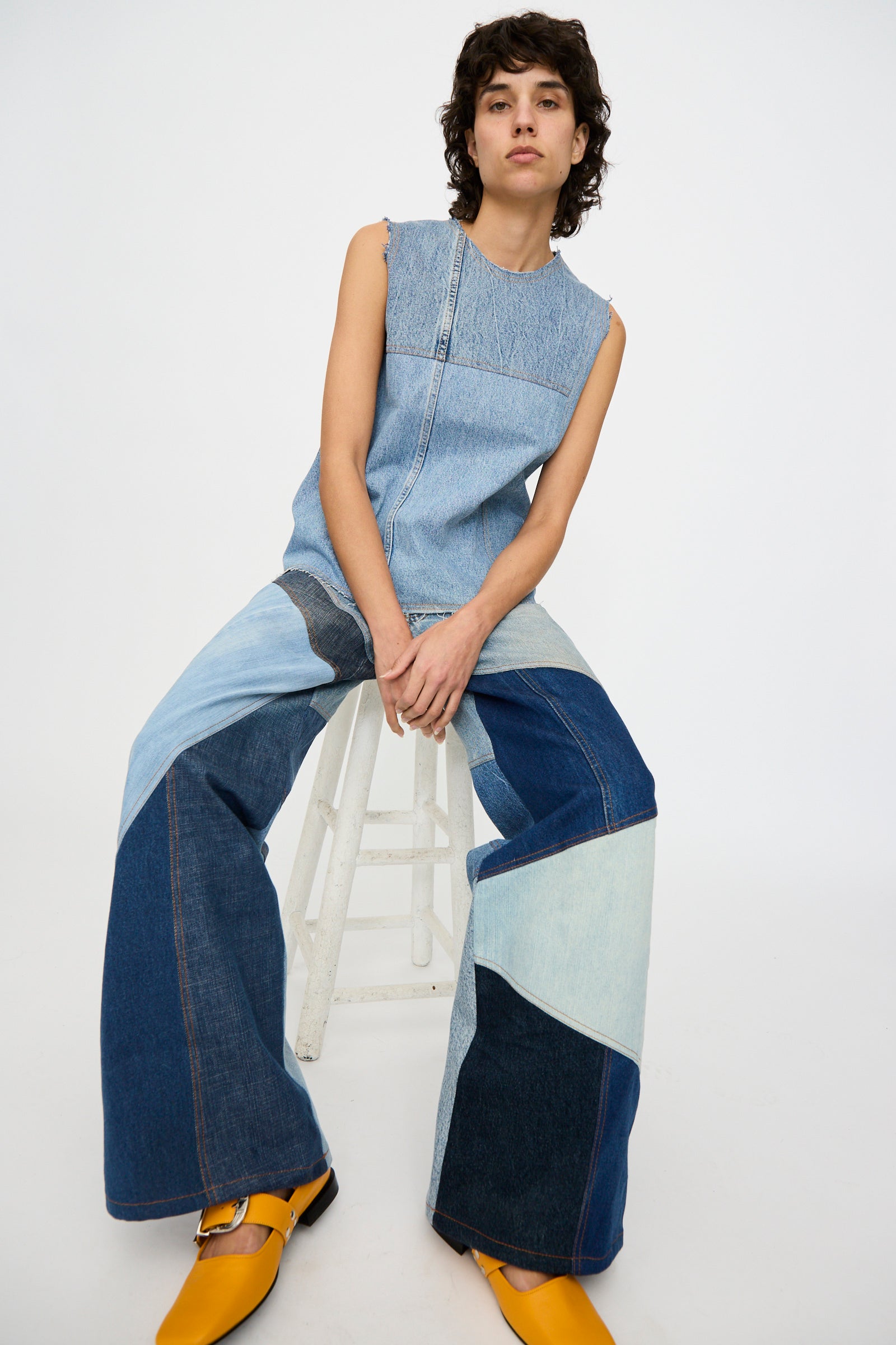 A person sits on a white stool against a plain background, wearing the Niccolò Pasqualetti Salvaged Jean Patchwork Carta Tank in Blue Denim with wide-leg patchwork jeans and yellow shoes.