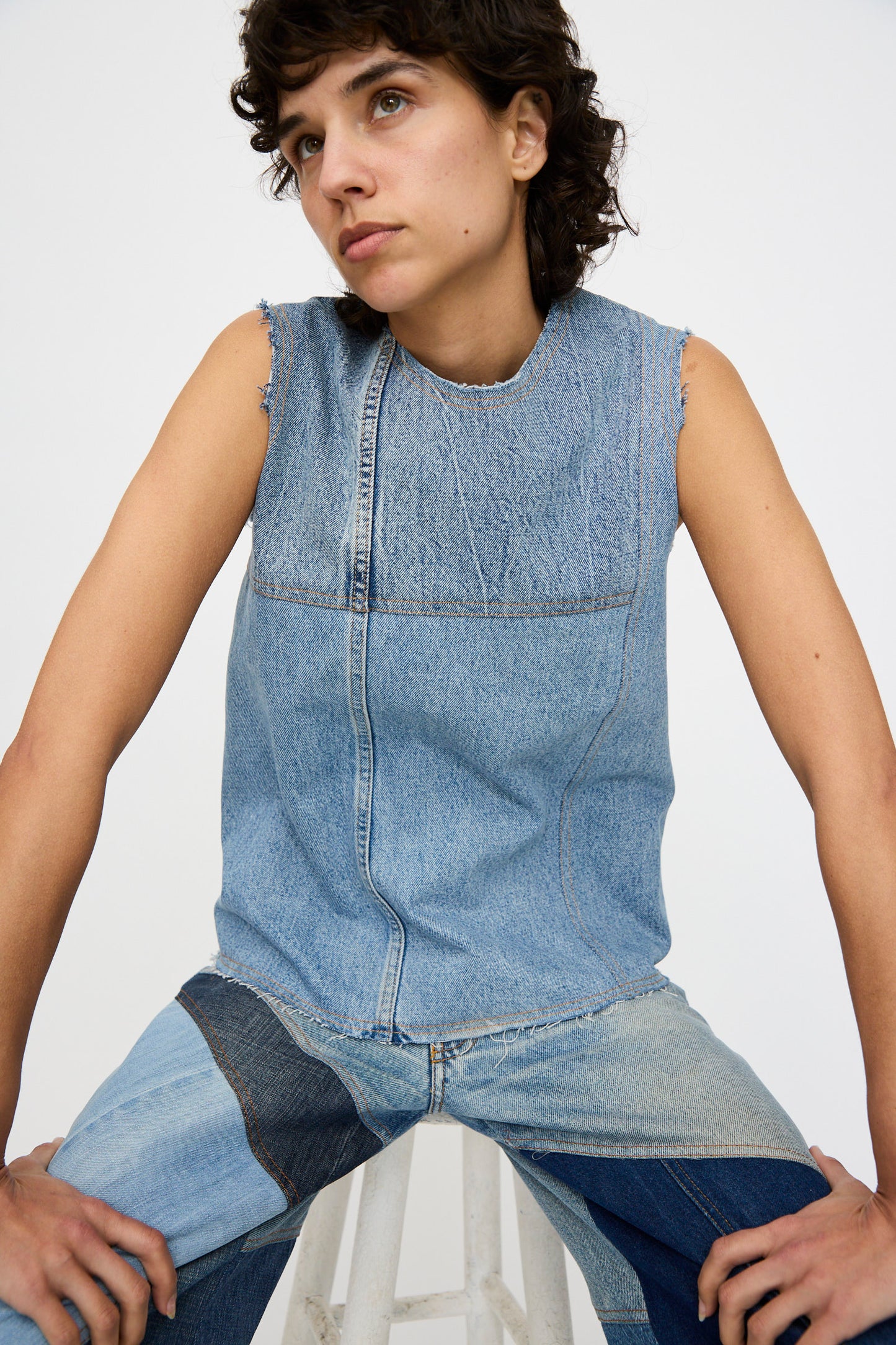 A person with short, curly hair sits on a stool against a white background wearing the Niccolò Pasqualetti Salvaged Jean Patchwork Carta Tank in Blue Denim.