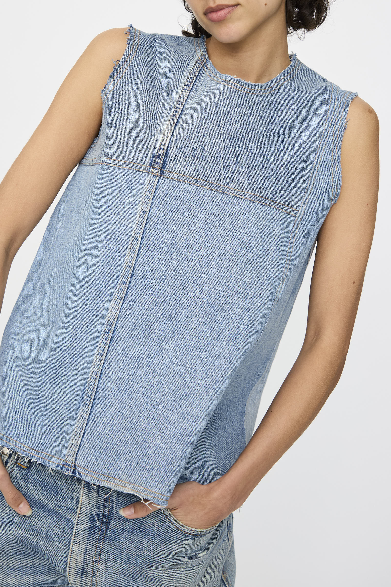 Model wears the Niccolò Pasqualetti Salvaged Jean Patchwork Carta Tank in Blue Denim, featuring raw edge hems, paired with light blue jeans; shown from shoulders to hips against a plain background.