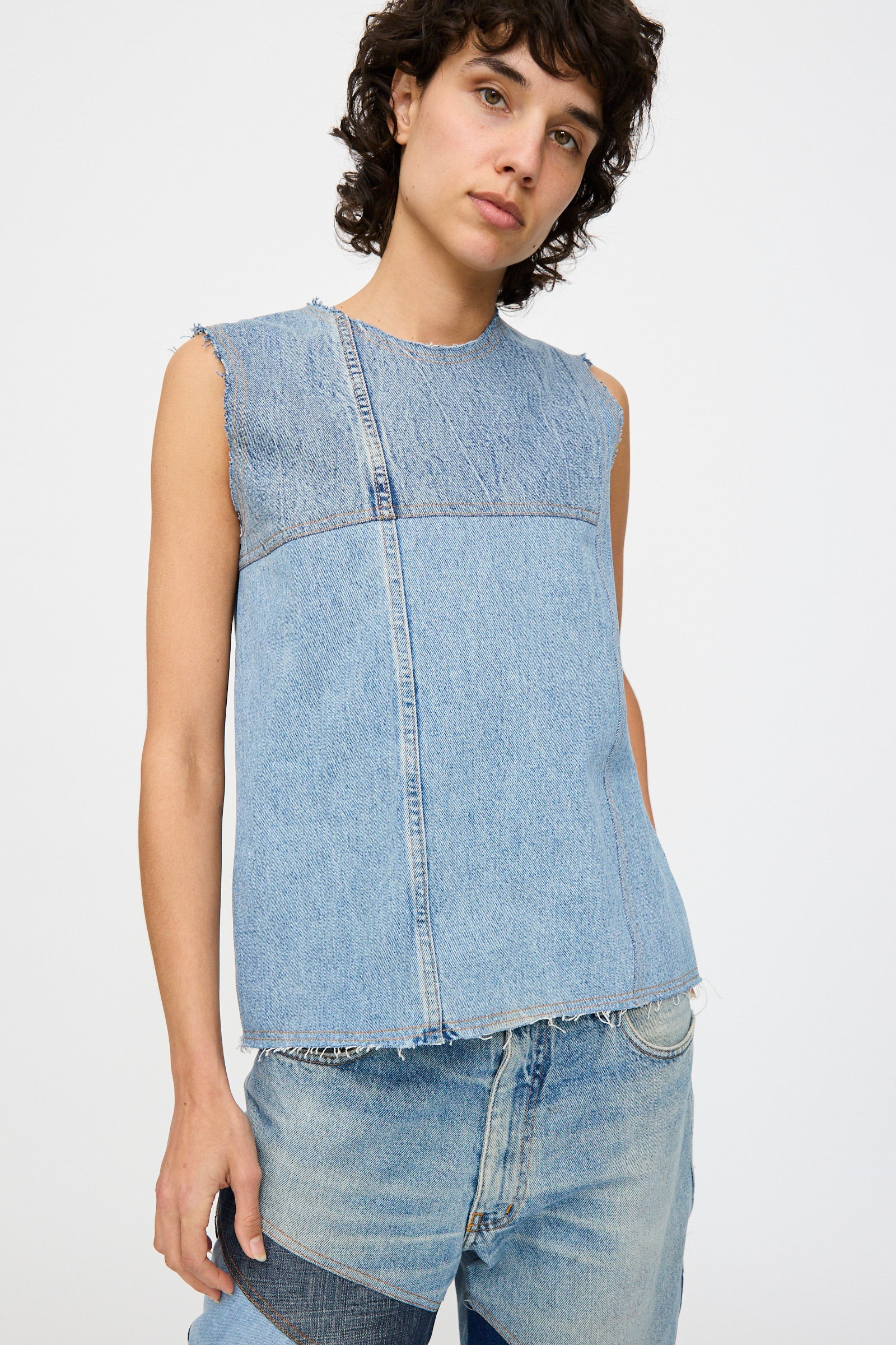 A person wears the Niccolò Pasqualetti Salvaged Jean Patchwork Carta Tank in Blue Denim, featuring raw edge hems, paired with light blue jeans and standing against a plain white background.