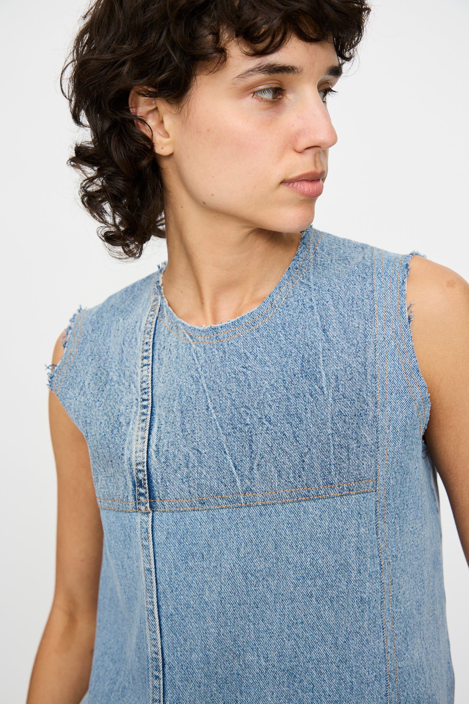 A person with short curly hair wears the Niccolò Pasqualetti Salvaged Jean Patchwork Carta Tank in Blue Denim, featuring raw edge hems, looking to the side against a plain background.