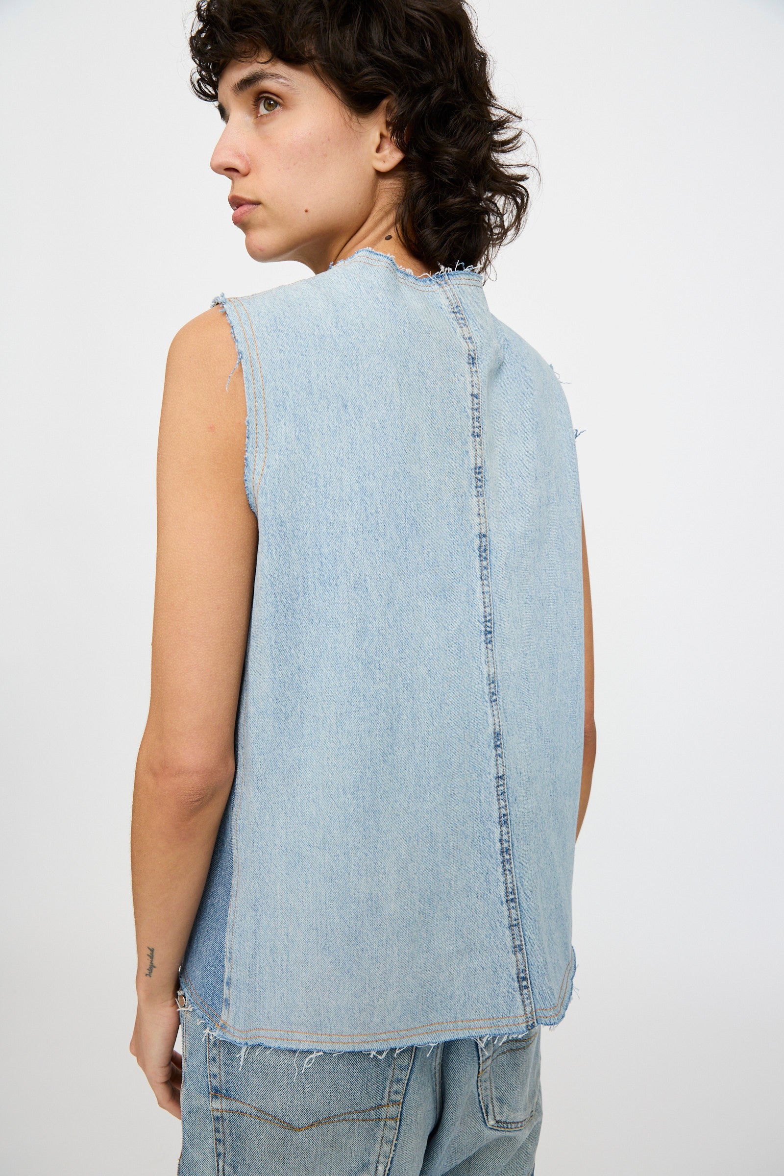 A person wearing the Niccolò Pasqualetti Salvaged Jean Patchwork Carta Tank in Blue Denim, with their back to the camera, styled with matching raw-edge hem denim shorts against a plain white background.