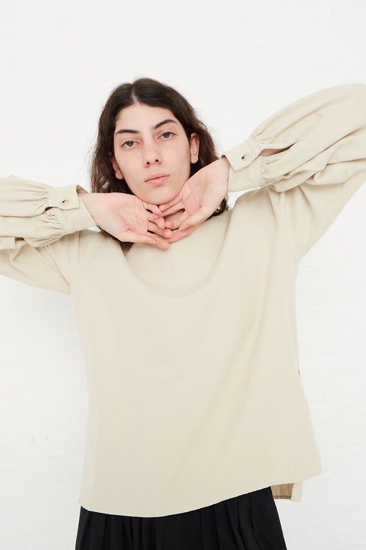 A woman wearing a beige Black Crane cotton twill long sleeve blouse with puffed sleeves. Front view. Model's arms are raised and placed under chin.