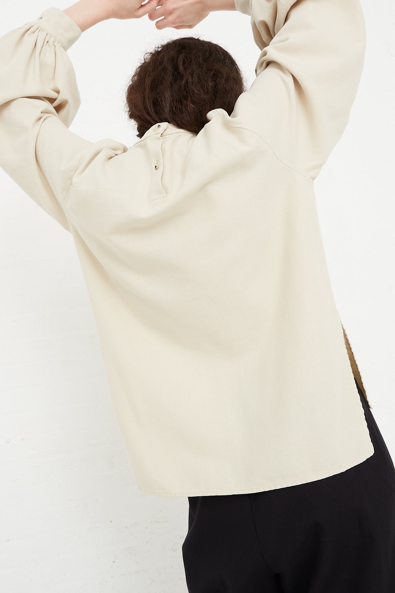 The model is wearing a Black Crane Cotton Twill Puff Sleeve Blouse in Ivory with long sleeves. Back view. Model's arms are raised above head.