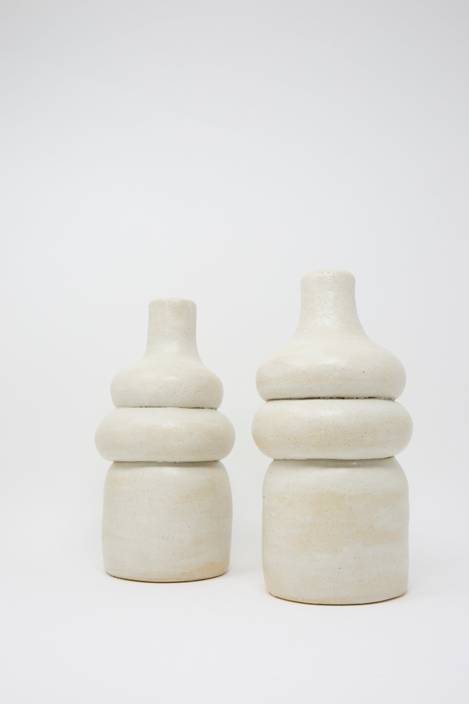 Two Prune Vessels by Oatmeal, featuring a white ceramic design with stacked cylindrical shapes, stand side by side against a plain background.