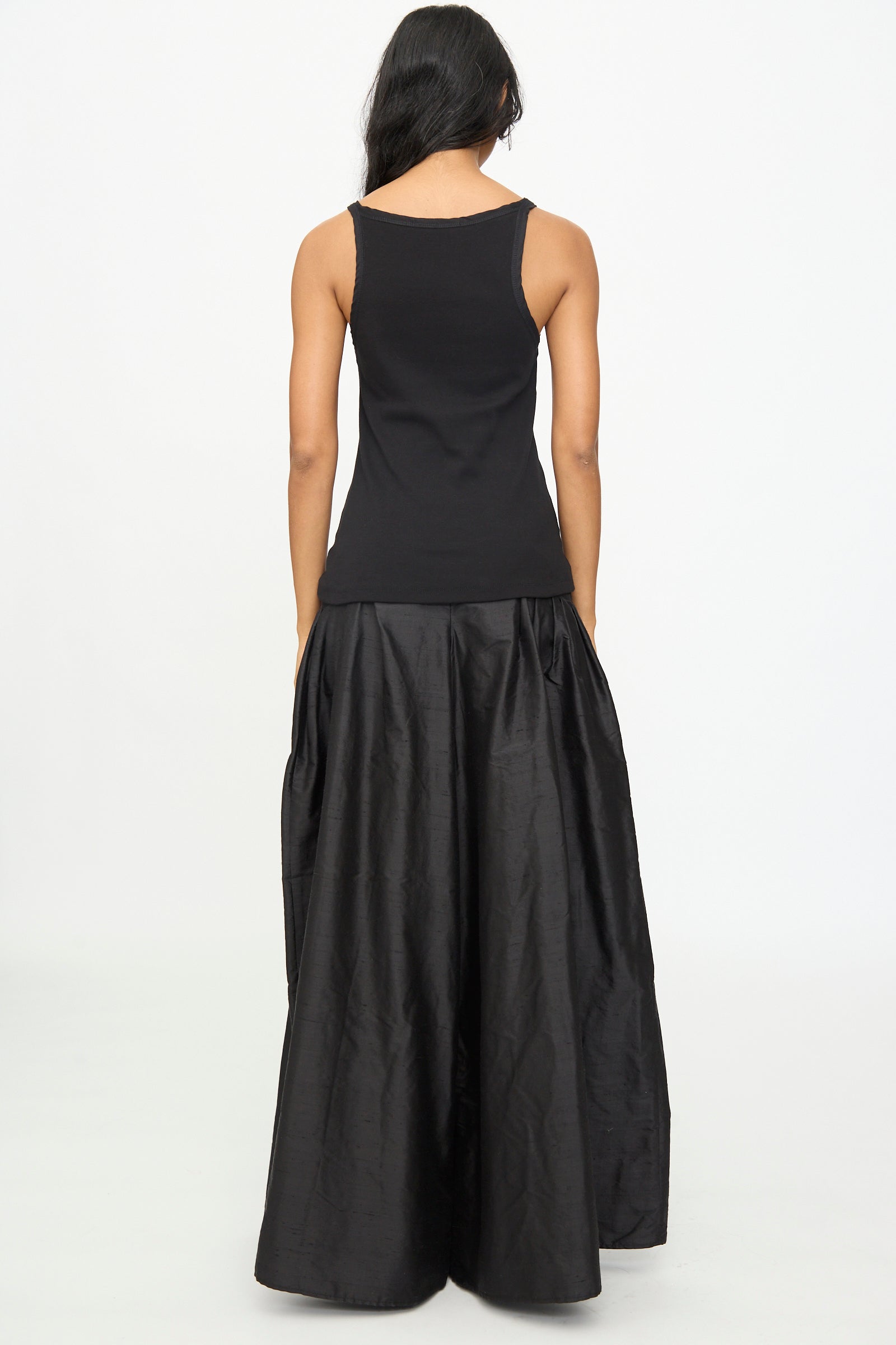 A woman facing away, wearing the Olivia Cashmore Billie Singlet in Black paired with a long, flowing black skirt, set against a plain white background.