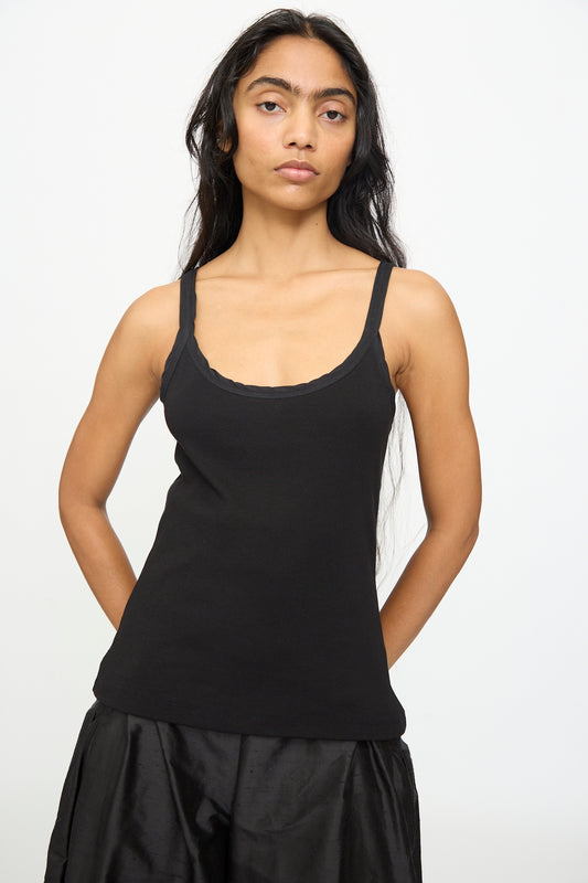 A person with long dark hair wears the Olivia Cashmore Billie Singlet in Black and a black skirt, standing in front of a plain white background with their arms behind their back.