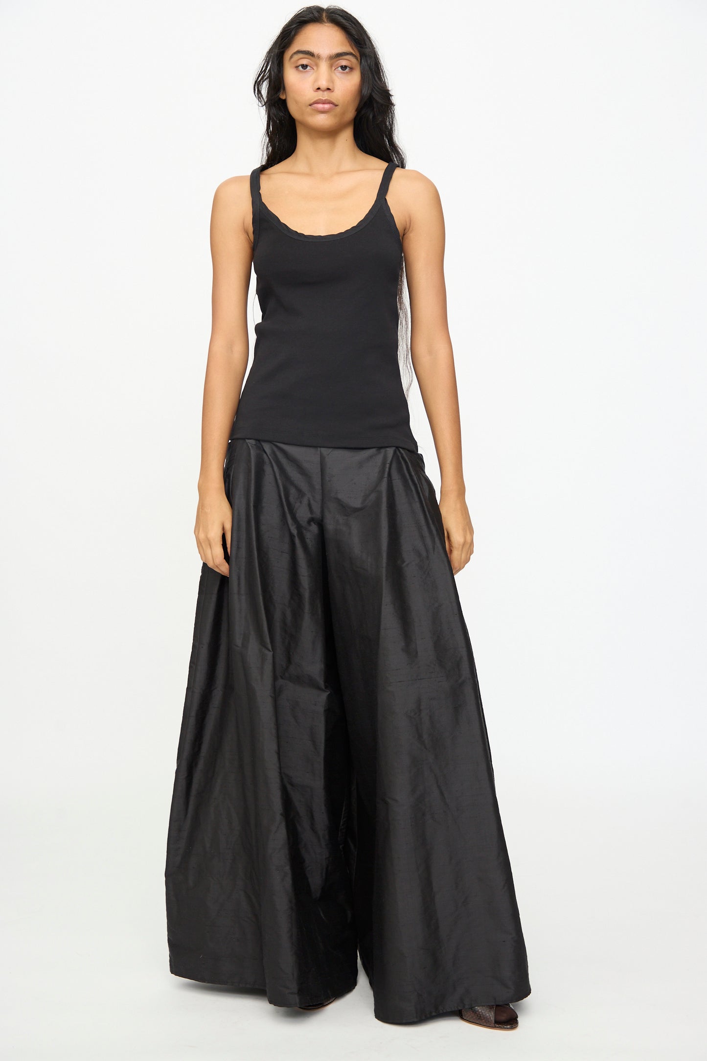 A person stands against a plain white background, wearing the Olivia Cashmore Billie Singlet in Black and wide-leg black pants, looking straight at the camera with a neutral expression.