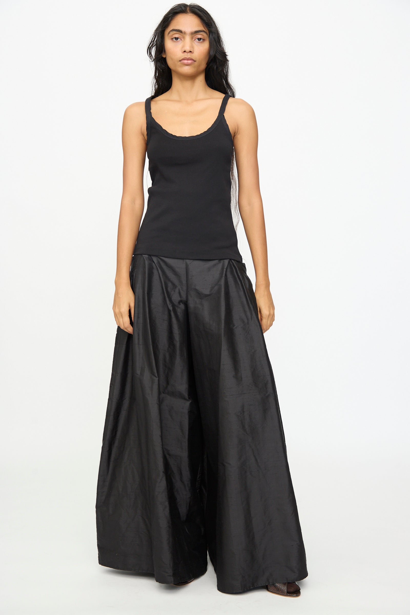 A person stands against a plain white background, wearing the Olivia Cashmore Billie Singlet in Black and wide-leg black pants, looking straight at the camera with a neutral expression.