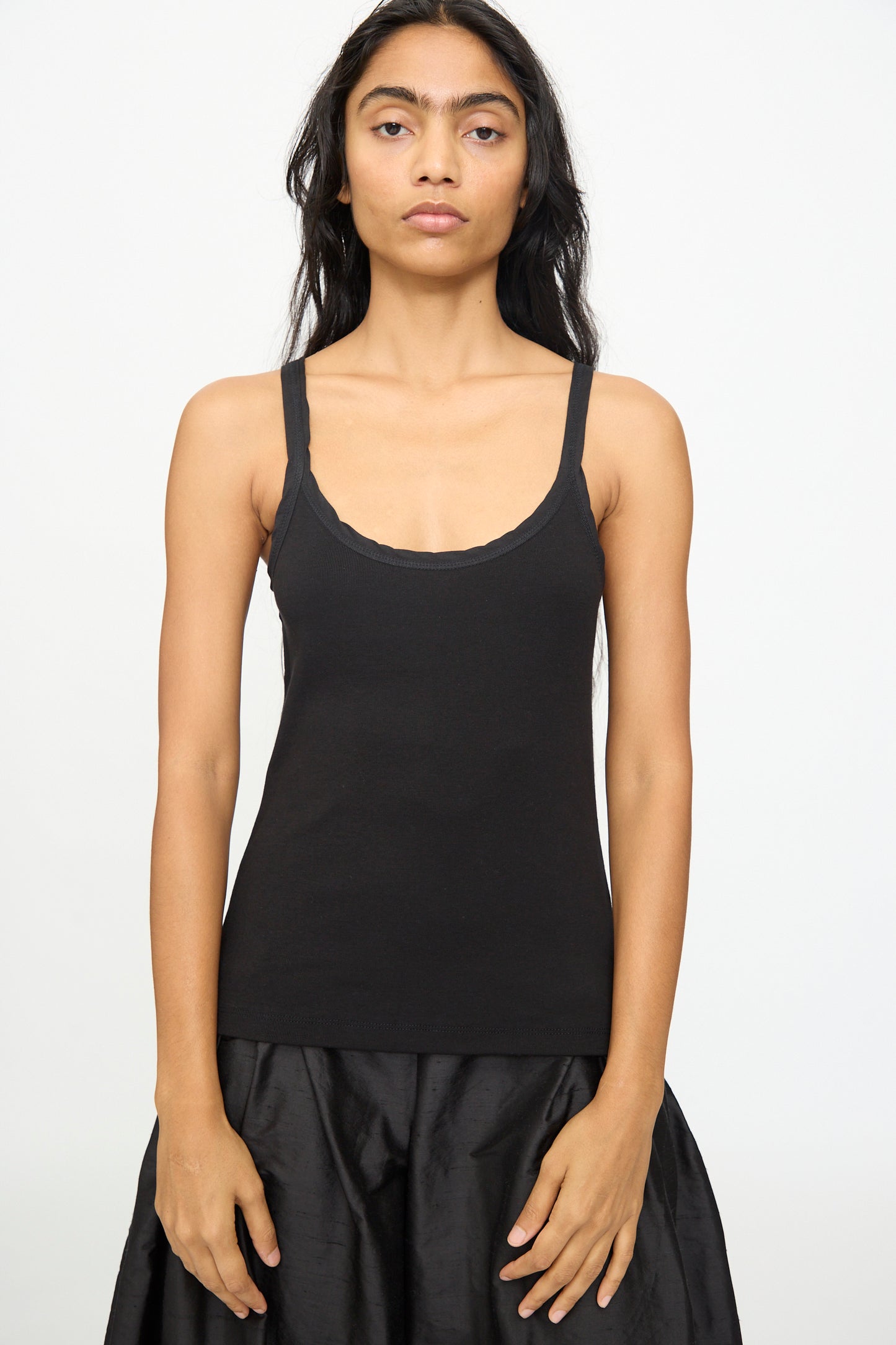 A person with long dark hair wears the Billie Singlet in Black by Olivia Cashmore and a black skirt, standing against a plain white background, facing the camera.