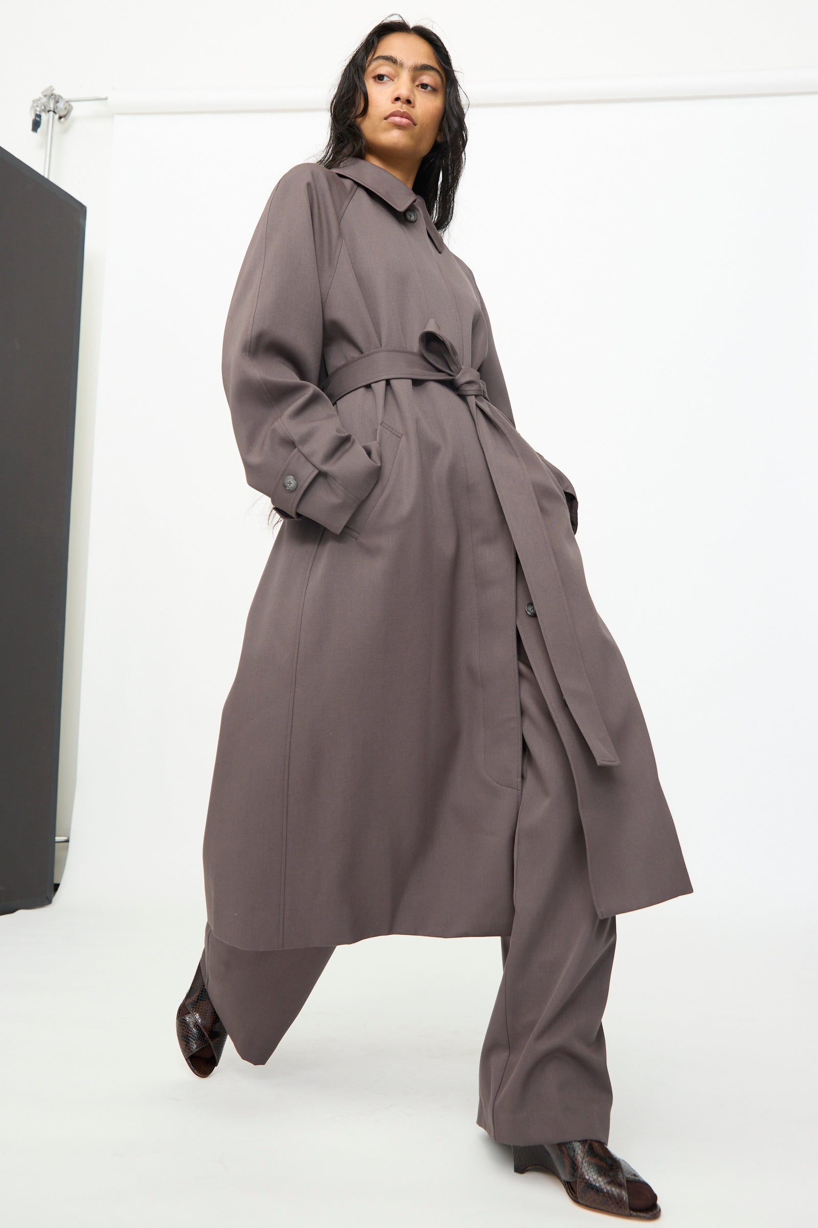Wearing the Gordie Coat in Mud by Olivia Cashmore, a person stands with one foot forward against a plain white background, styled in matching wide-leg pants.