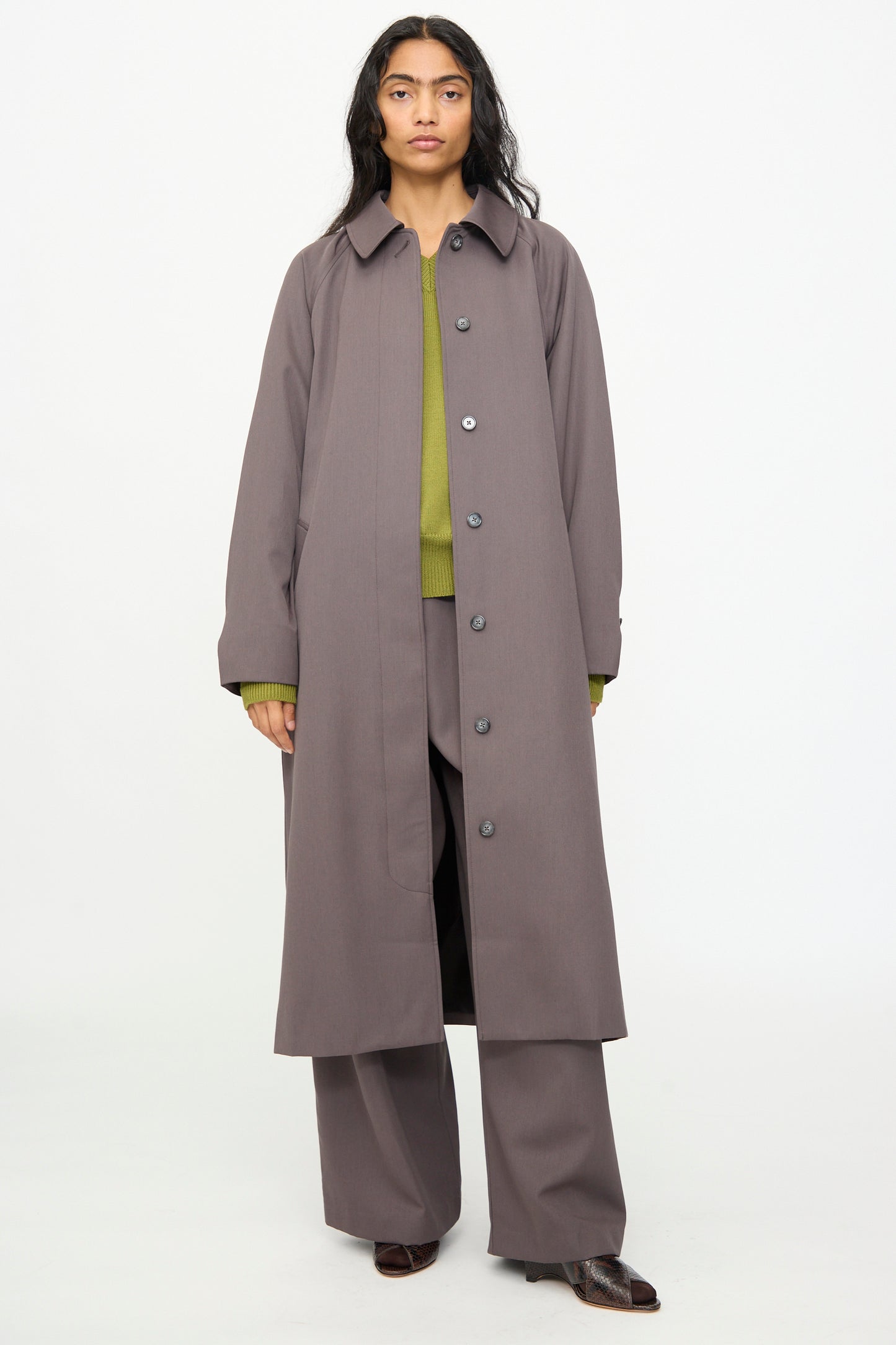 A person stands facing forward, wearing the Olivia Cashmore Gordie Coat in Mud—an Italian wool coat made in New Zealand—styled over wide-leg gray pants and a green sweater with brown shoes, against a plain white background.