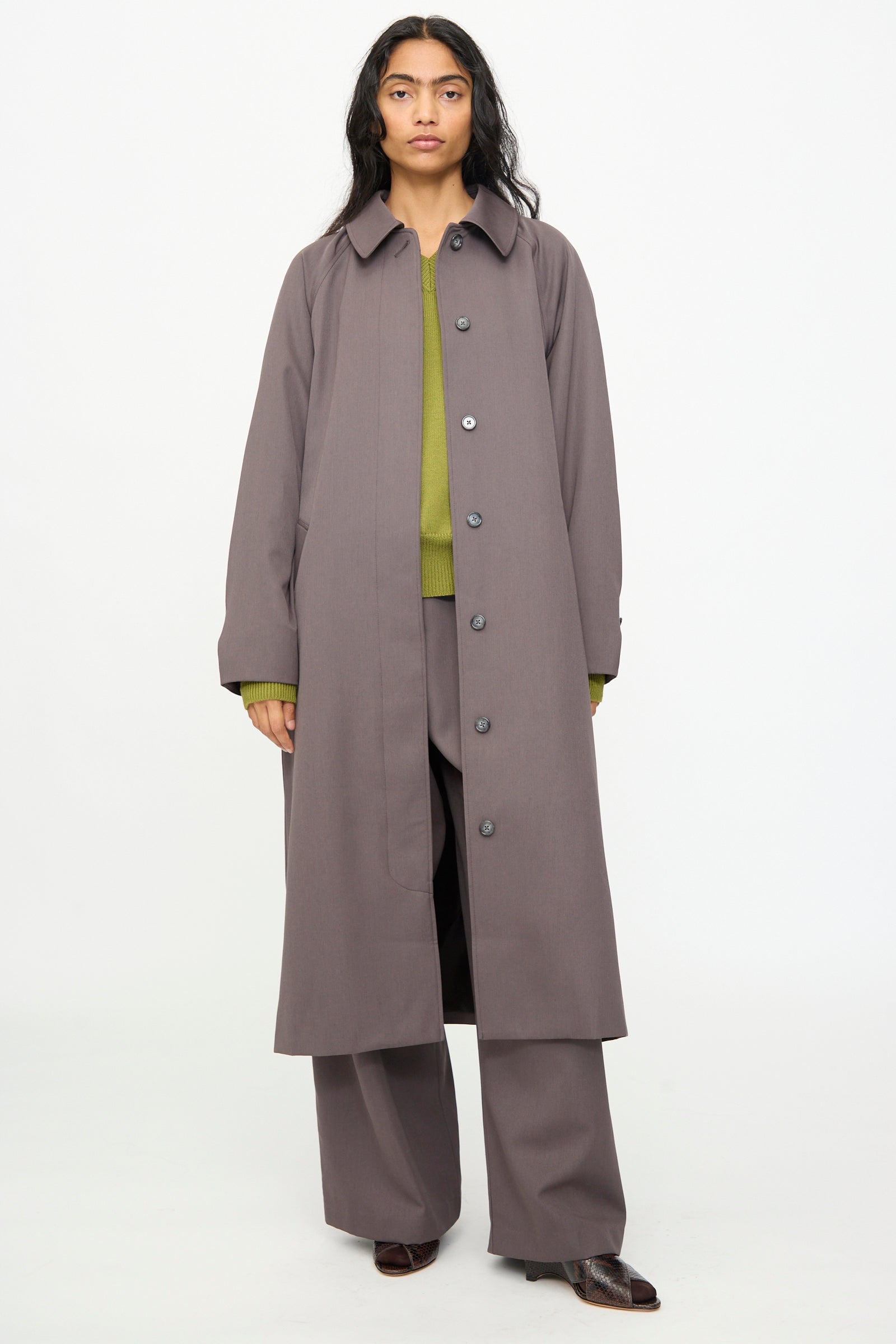 A person stands facing forward, wearing the Olivia Cashmore Gordie Coat in Mud—an Italian wool coat made in New Zealand—styled over wide-leg gray pants and a green sweater with brown shoes, against a plain white background.