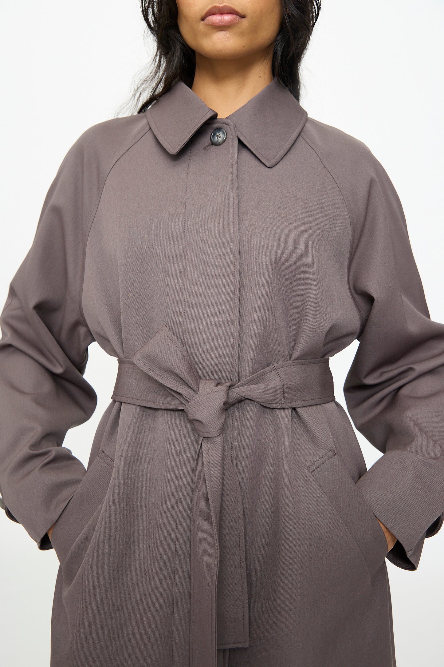 A person wearing the Gordie Coat in Mud by Olivia Cashmore, a relaxed Italian wool coat featuring a collar, hands in pockets, with the top button fastened.