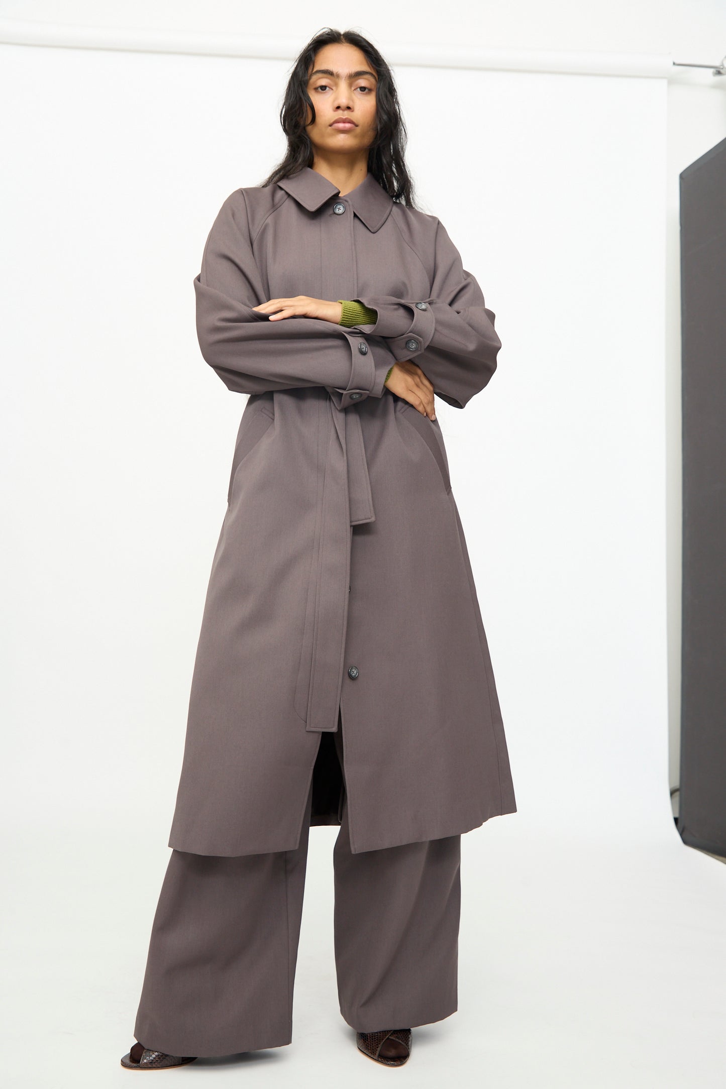 A person stands in a studio wearing the Olivia Cashmore Gordie Coat in Mud over wide-leg pants and dark shoes, looking directly at the camera.