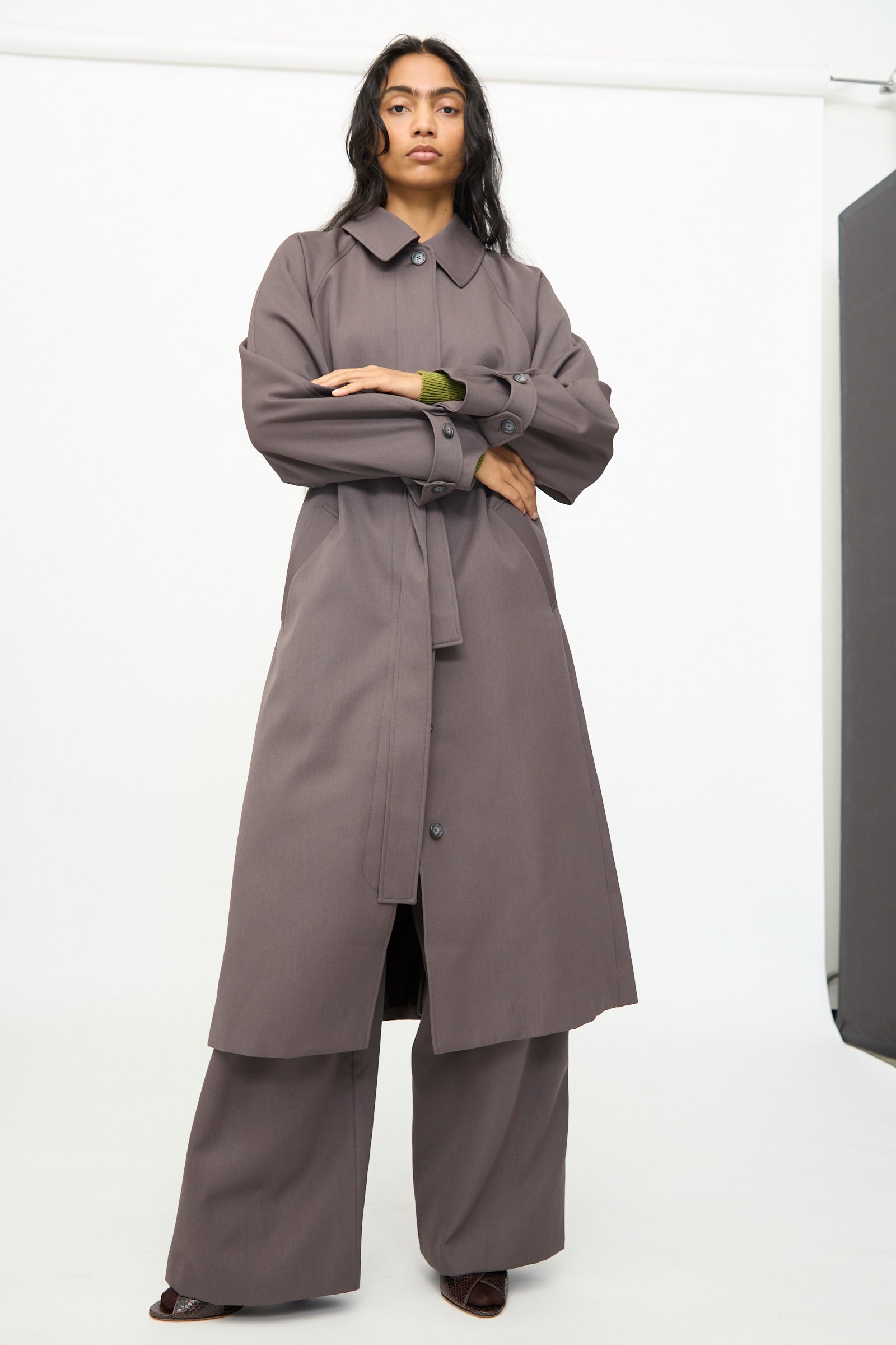 A person stands in a studio wearing the Olivia Cashmore Gordie Coat in Mud over wide-leg pants and dark shoes, looking directly at the camera.