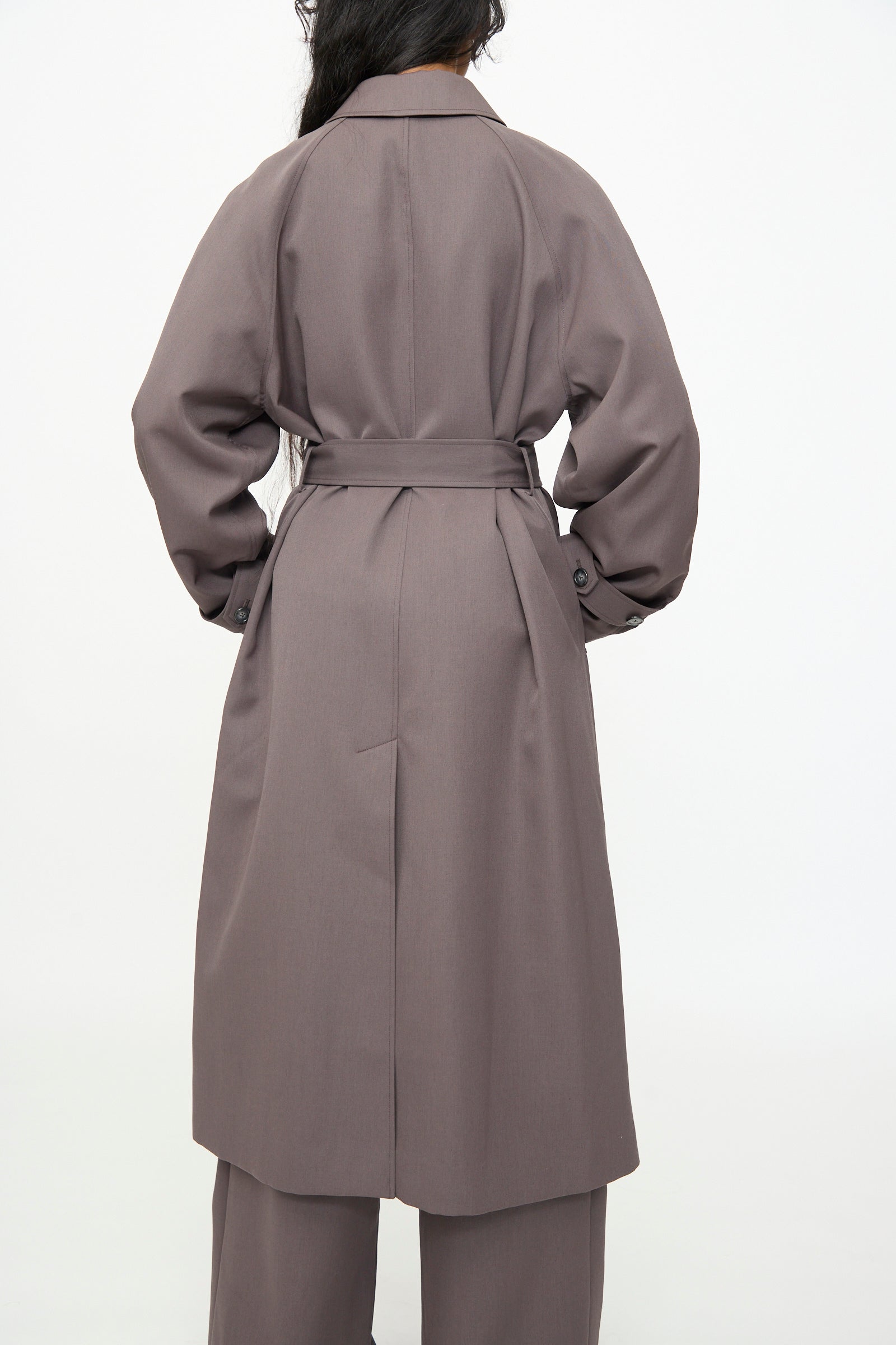 A person with long dark hair is photographed from the back against a plain white background, wearing the Olivia Cashmore Gordie Coat in Mud with matching pants.