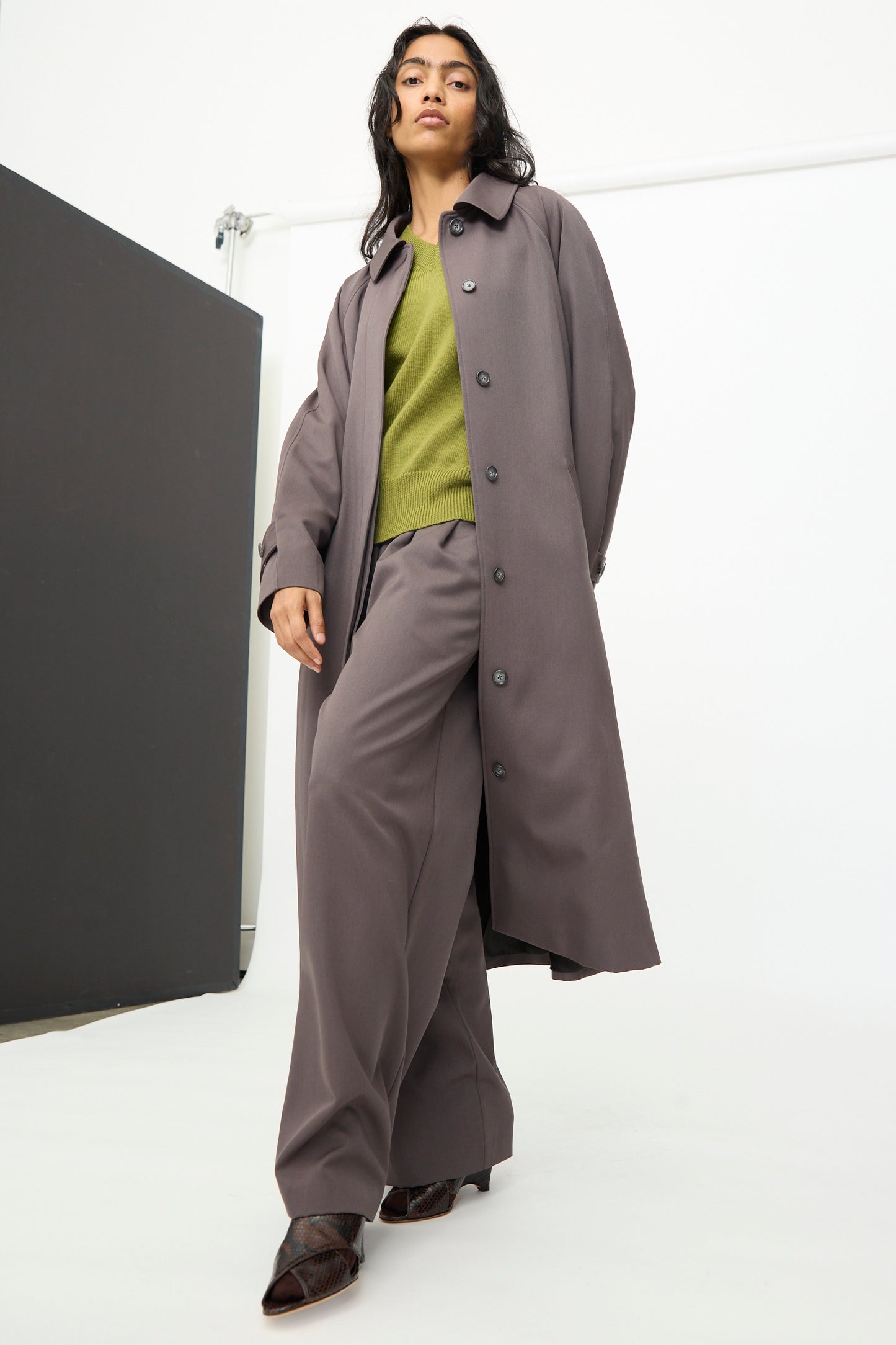 A person stands in a minimalist studio setting wearing the Olivia Cashmore Gordie Coat in Mud, styled as a relaxed coat and paired with wide-leg pants, a green sweater, and brown shoes.