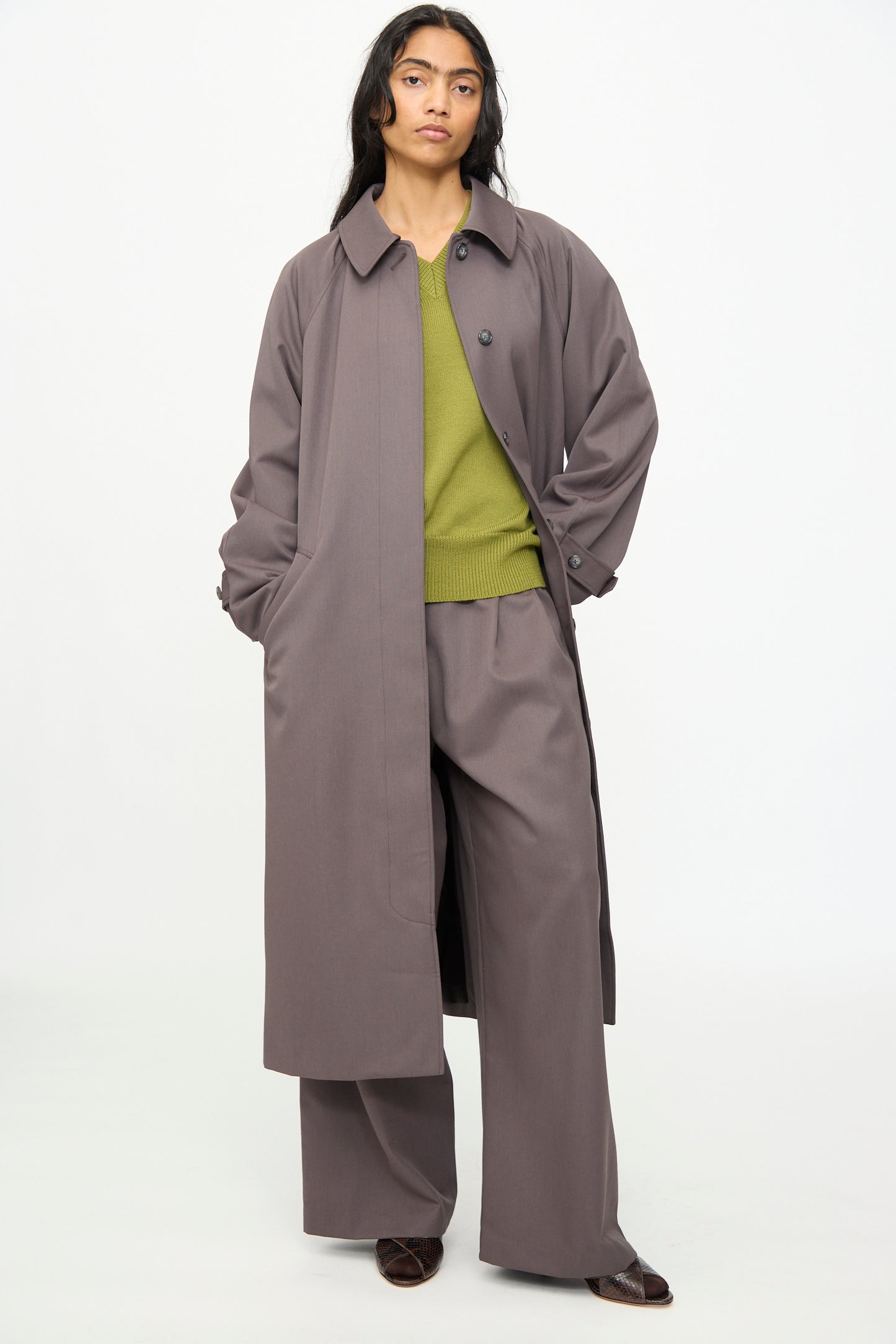 Wearing the Olivia Cashmore Gordie Coat in Mud, paired with wide-legged gray trousers and an olive green sweater, a person stands against a plain white background.