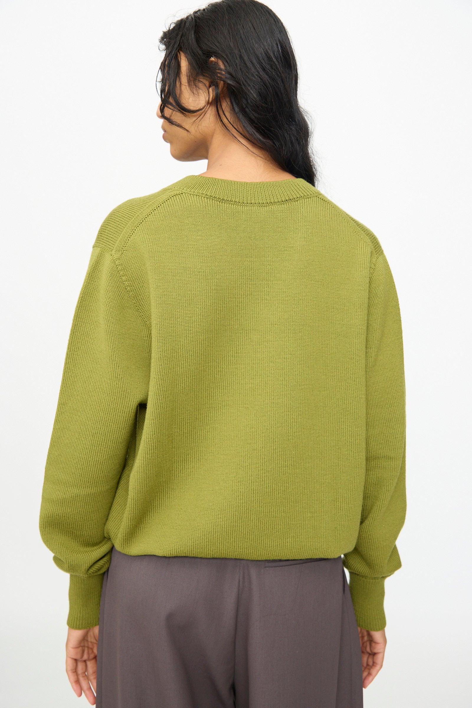 A person with long dark hair is shown from the back against a plain light background, wearing the Olivia Cashmore Merino Poppy Knit Sweater in Pickle and gray pants.