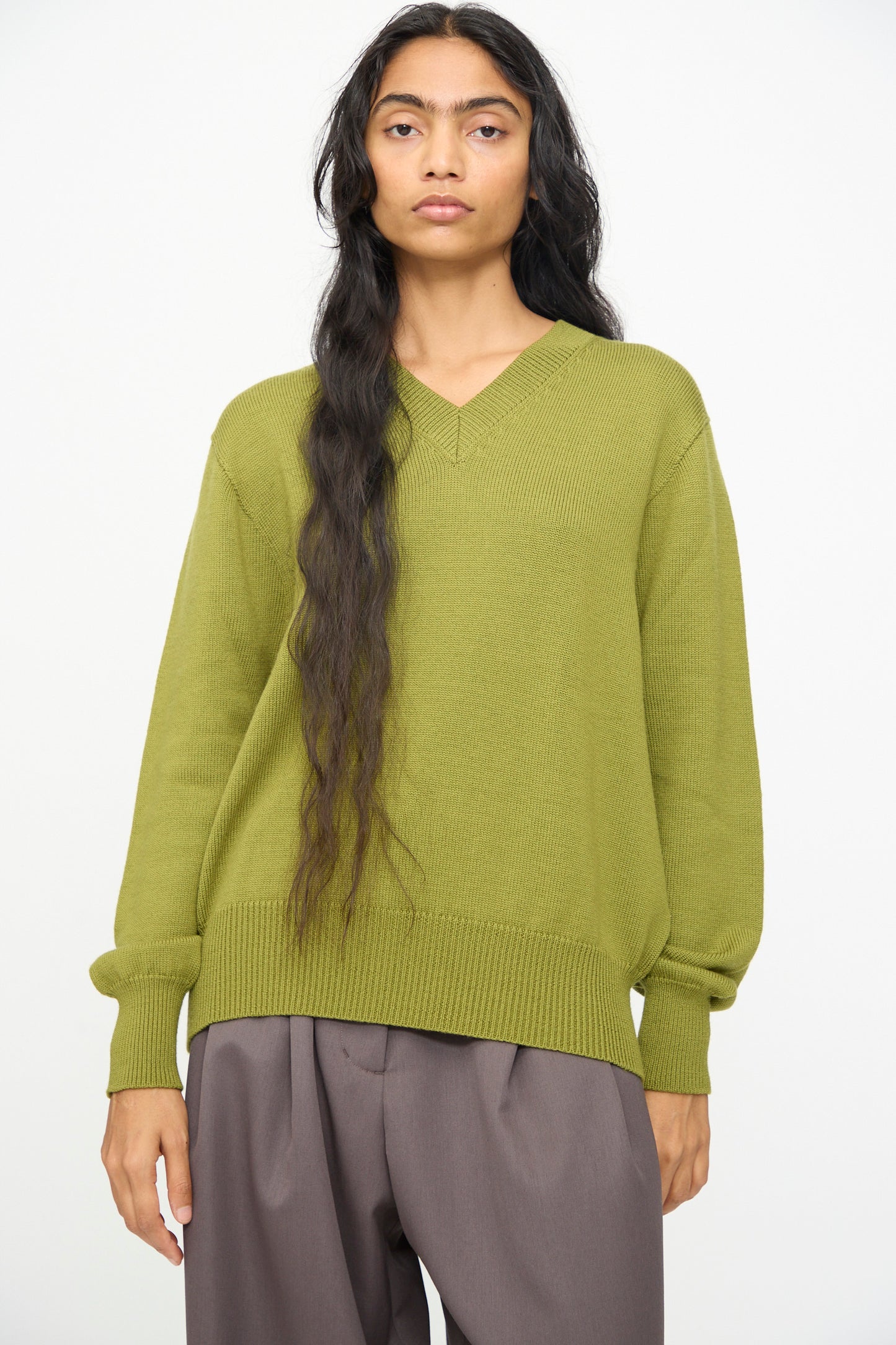Wearing the Olivia Cashmore Merino Poppy Knit Sweater in Pickle, a person with long dark hair and loose brown pants stands against a plain light background.