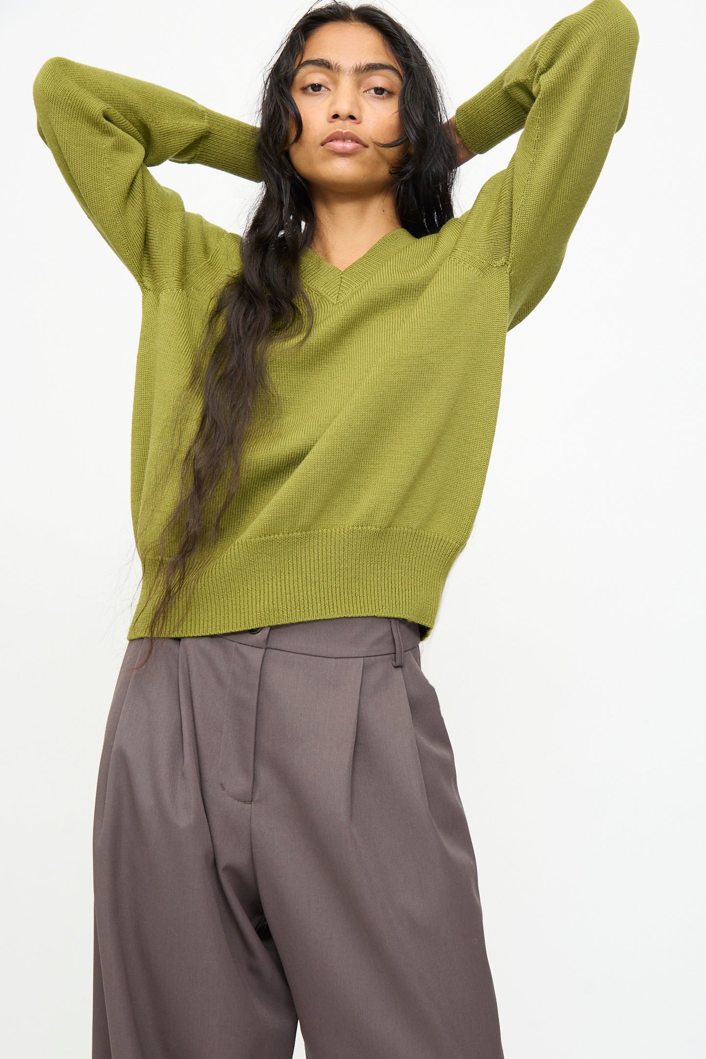 A person wearing the Olivia Cashmore Merino Poppy Knit Sweater in Pickle and loose brown trousers stands with their hands resting behind their head against a plain light background.