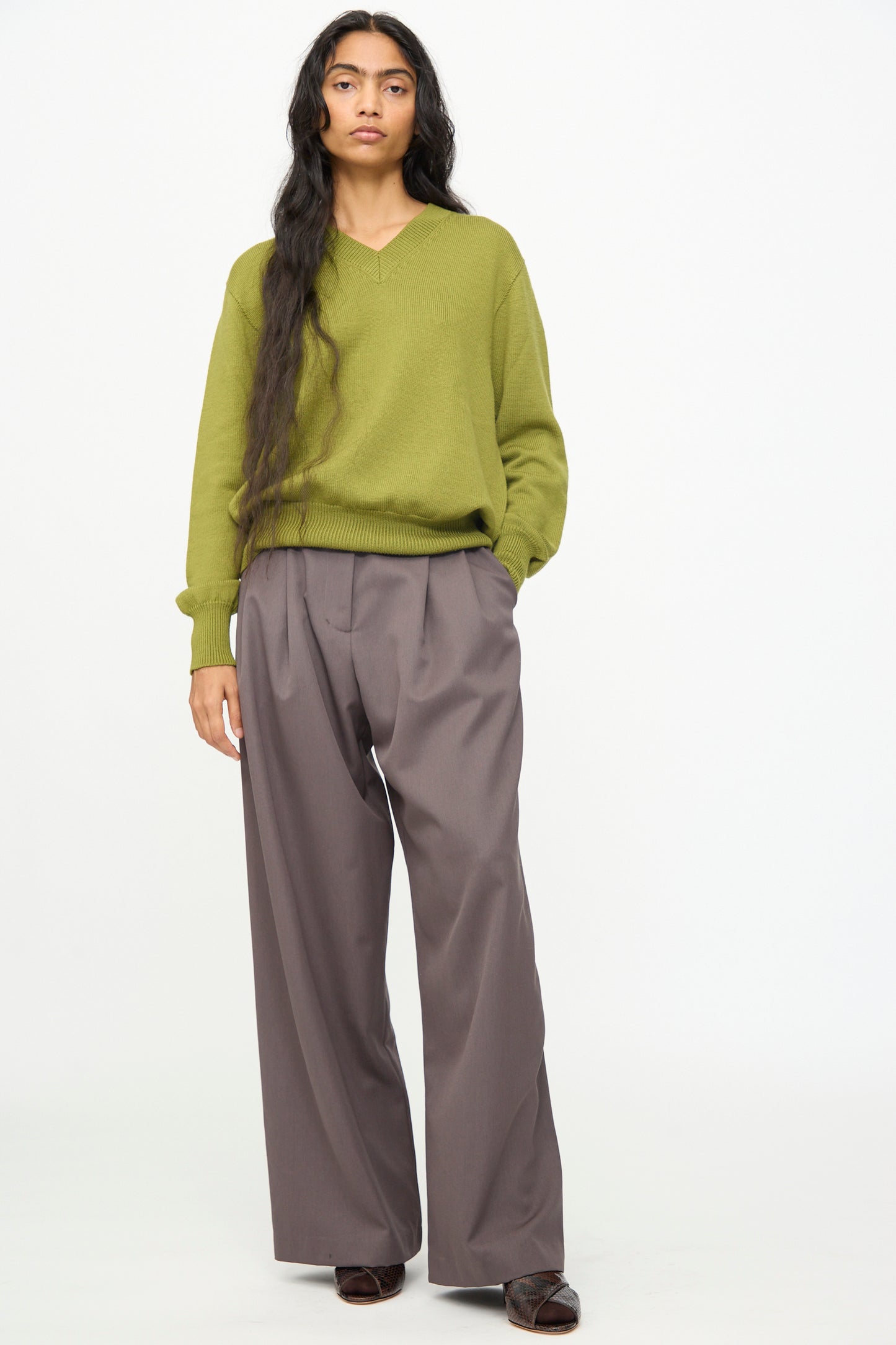 A person stands against a plain background wearing the Olivia Cashmore Merino Poppy Knit Sweater in Pickle, paired with loose-fitting gray trousers and dark shoes, their long dark hair draped over one shoulder.