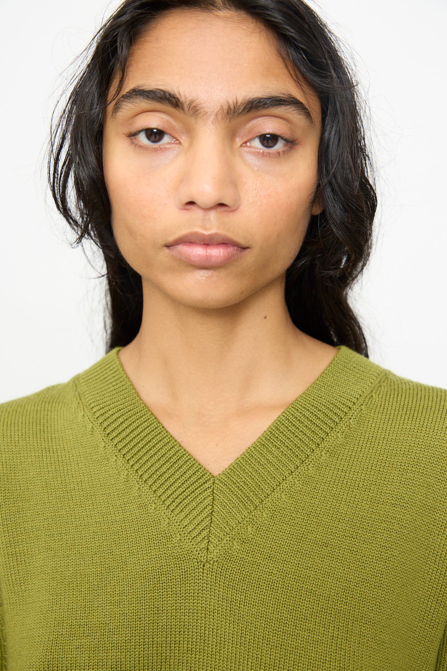 A person with long dark hair is wearing the Olivia Cashmore Merino Poppy Knit Sweater in Pickle—a green merino wool V-neck style made in New Zealand—facing forward with a neutral expression against a plain background.