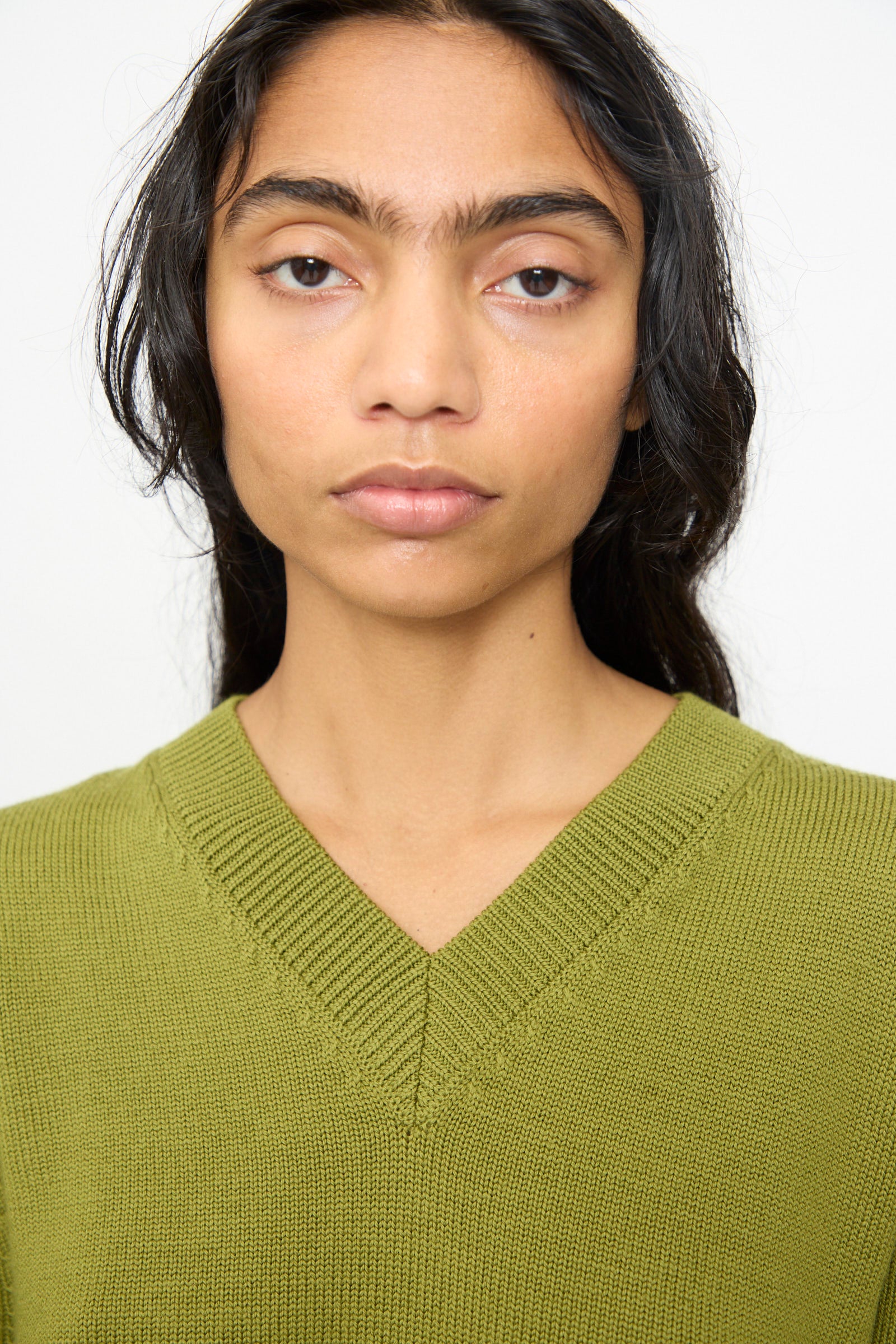 A person with long dark hair is wearing the Olivia Cashmore Merino Poppy Knit Sweater in Pickle—a green merino wool V-neck style made in New Zealand—facing forward with a neutral expression against a plain background.