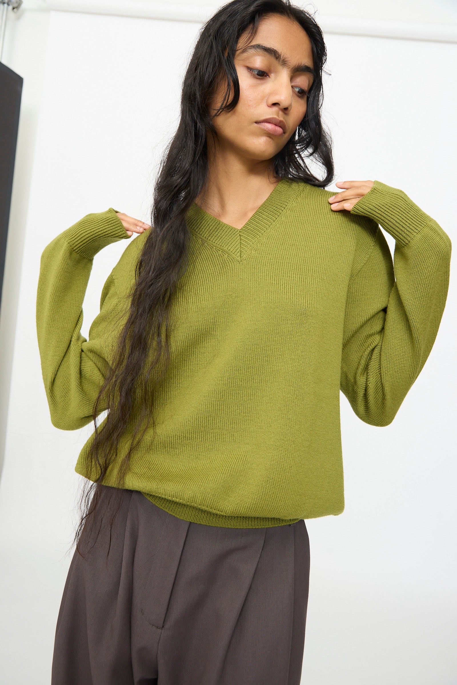 A person wearing the Olivia Cashmore Merino Poppy Knit Sweater in Pickle and brown pants stands with hands raised near their shoulders against a plain background. This stylish sweater is crafted from merino wool and made in New Zealand.