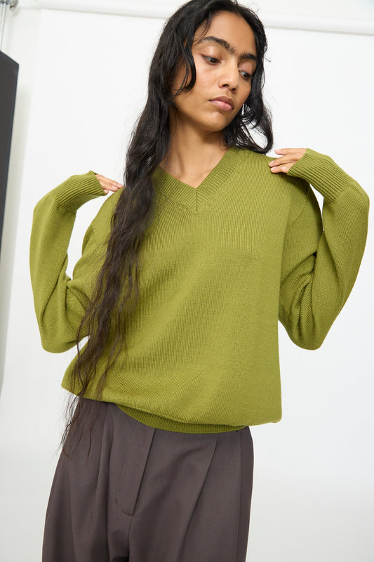 A person wearing the Olivia Cashmore Merino Poppy Knit Sweater in Pickle and brown pants stands with hands raised near their shoulders against a plain background. This stylish sweater is crafted from merino wool and made in New Zealand.