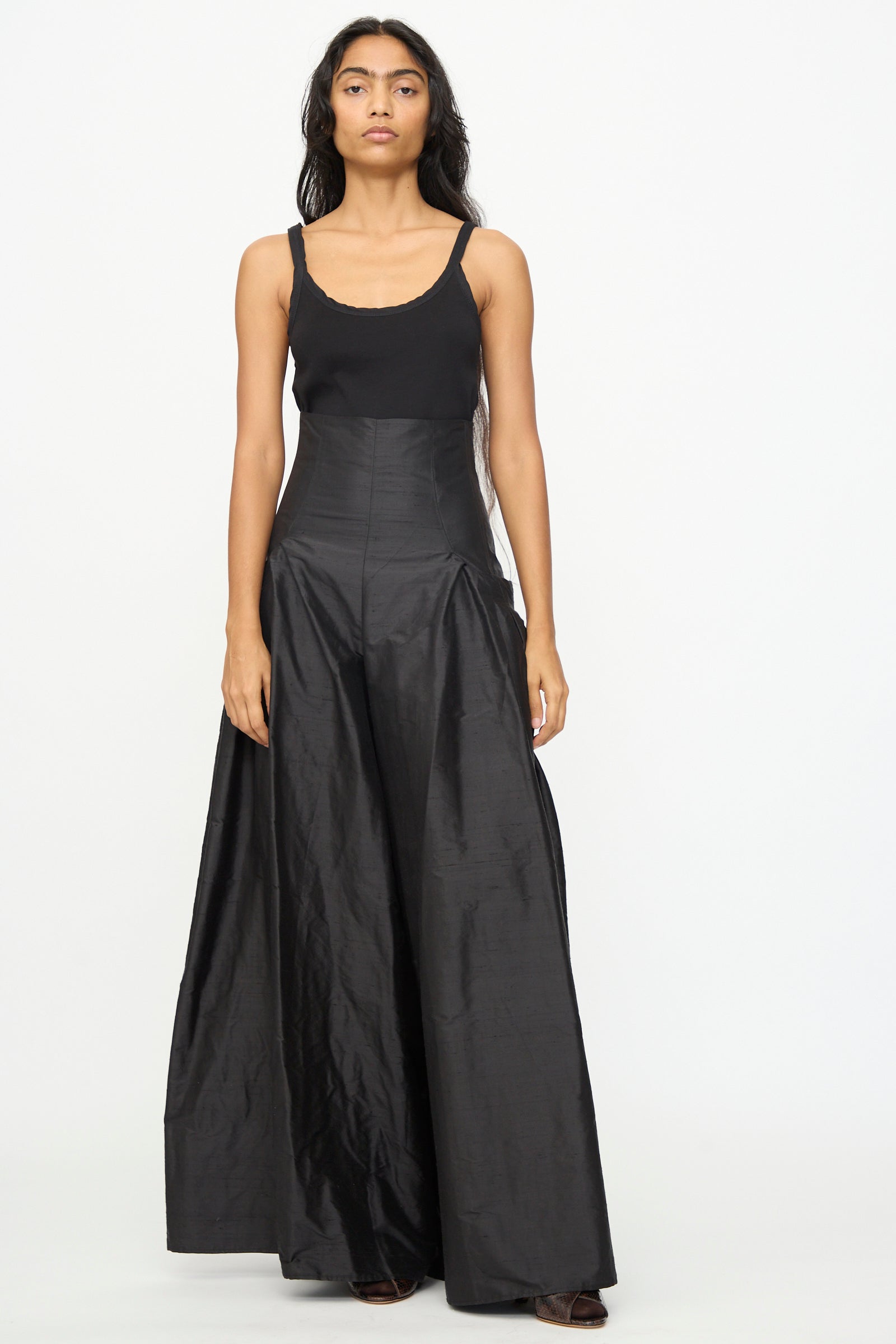 A woman stands facing forward wearing the Olivia Cashmore Robbie Pant in Black—high-waisted, wide-leg silk blend trousers—paired with a sleeveless black top against a plain white background.