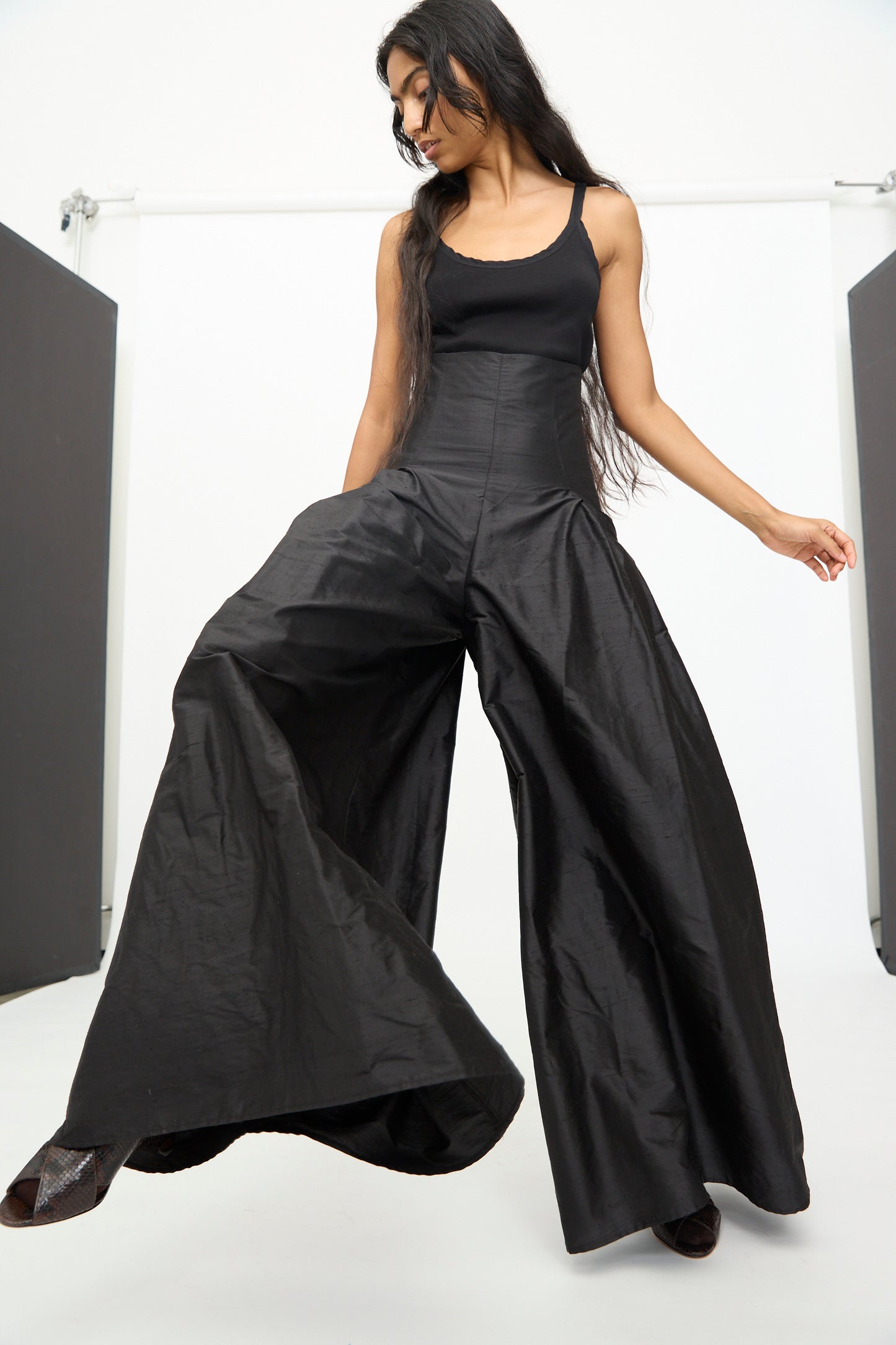 A person wearing the Olivia Cashmore Robbie Pant in Black stands indoors against a white background, looking down and slightly to the side.
