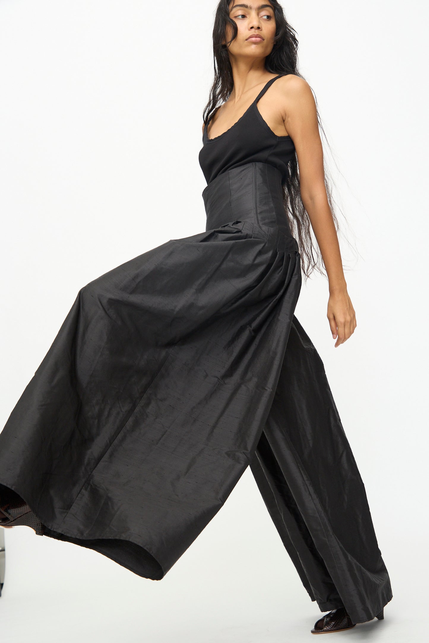 A woman wearing a sleeveless black top and the Olivia Cashmore Robbie Pant in Black—a chic, silk blend, wide-leg, high-waisted style—walks against a plain white background.