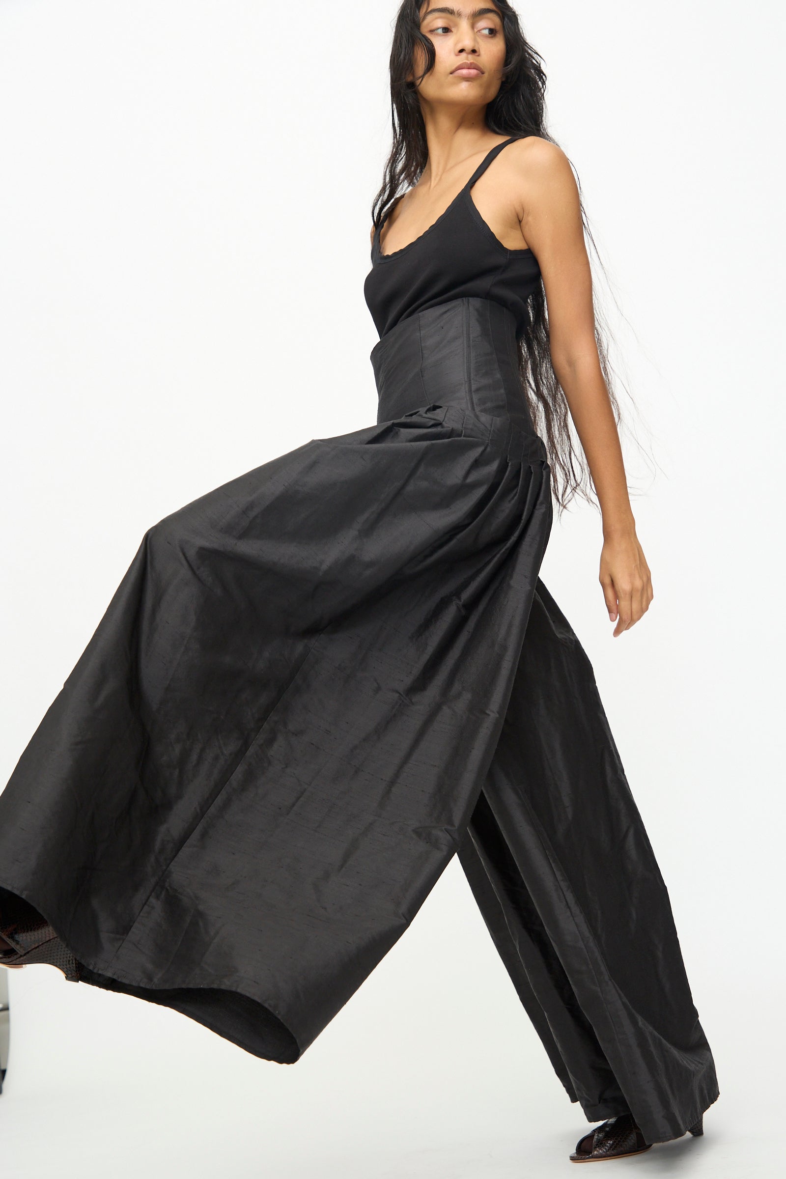 A woman wearing a sleeveless black top and the Olivia Cashmore Robbie Pant in Black—a chic, silk blend, wide-leg, high-waisted style—walks against a plain white background.