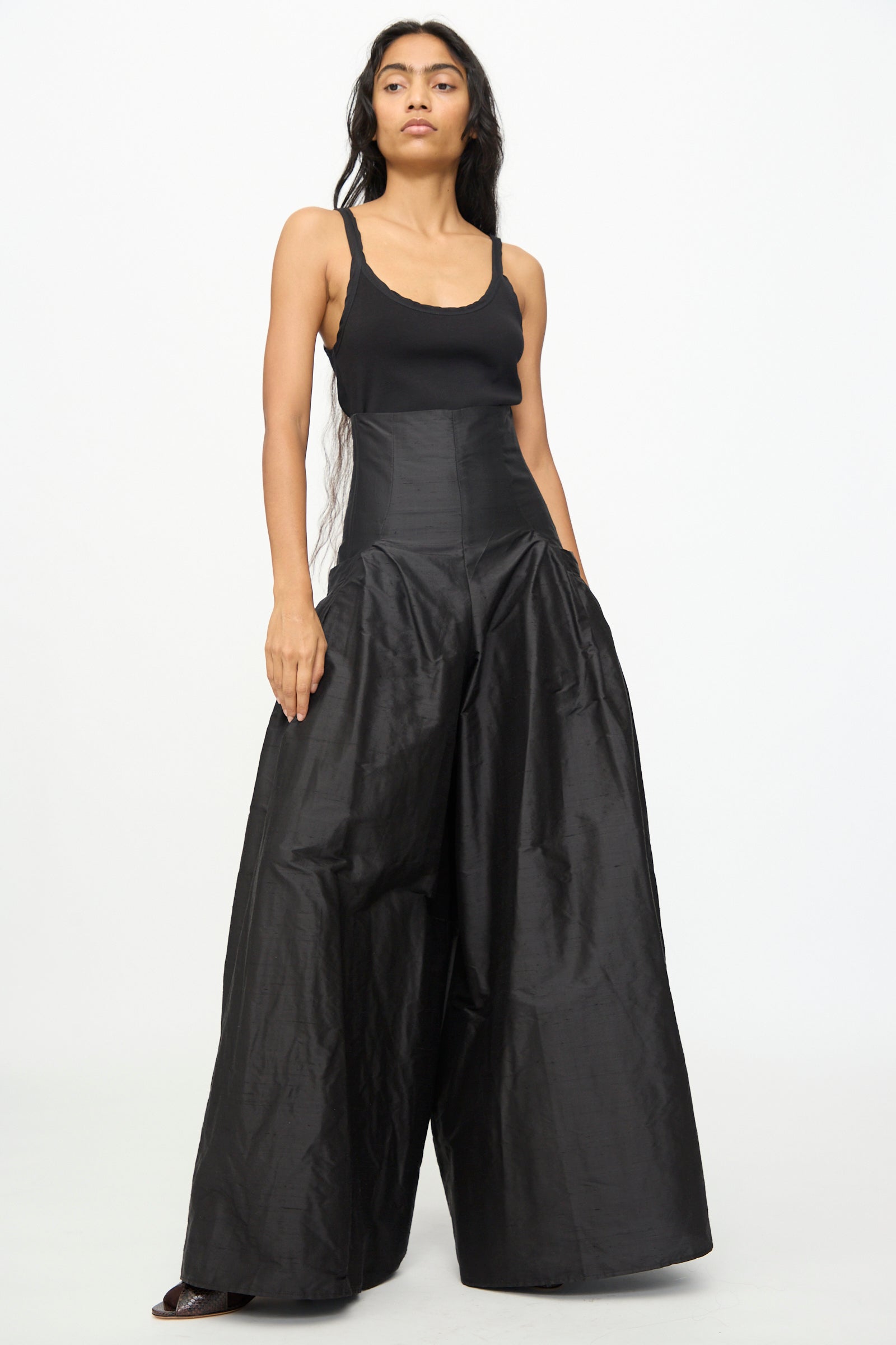 A woman stands against a white background, wearing the Olivia Cashmore Robbie Pant in Black—a high-waisted, wide leg style crafted from a luxurious silk blend—paired with a black tank top and her hands in her pockets.