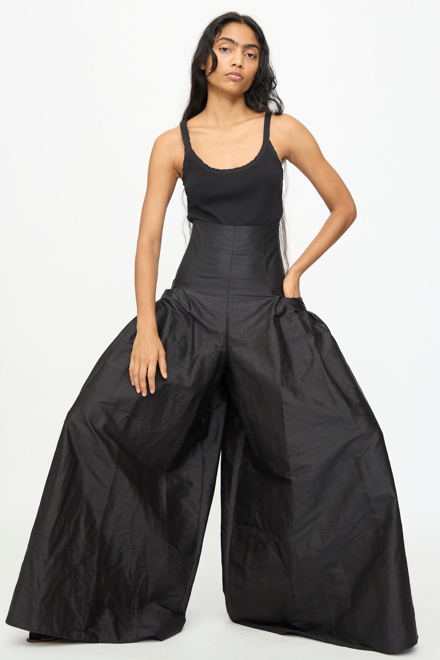 A person wearing a black sleeveless top and the Olivia Cashmore Robbie Pant in Black, featuring a high waist and voluminous wide legs, stands against a plain white background. Crafted from a refined silk blend, the pants add an elegant flow to the overall look.