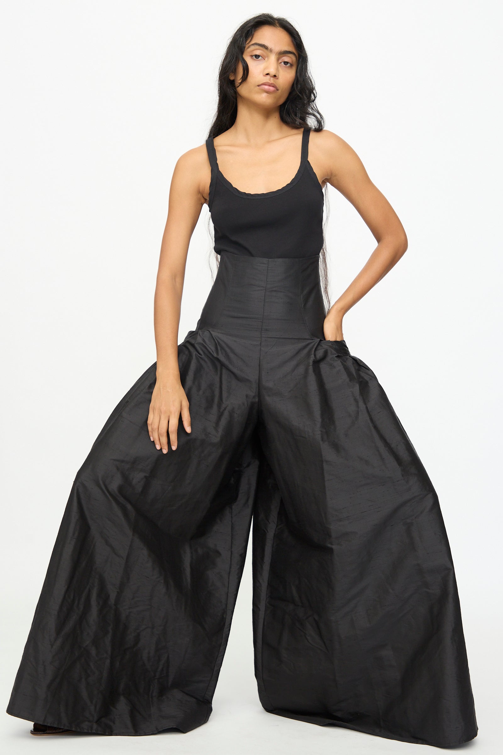 A person wearing a black sleeveless top and the Olivia Cashmore Robbie Pant in Black, featuring a high waist and voluminous wide legs, stands against a plain white background. Crafted from a refined silk blend, the pants add an elegant flow to the overall look.