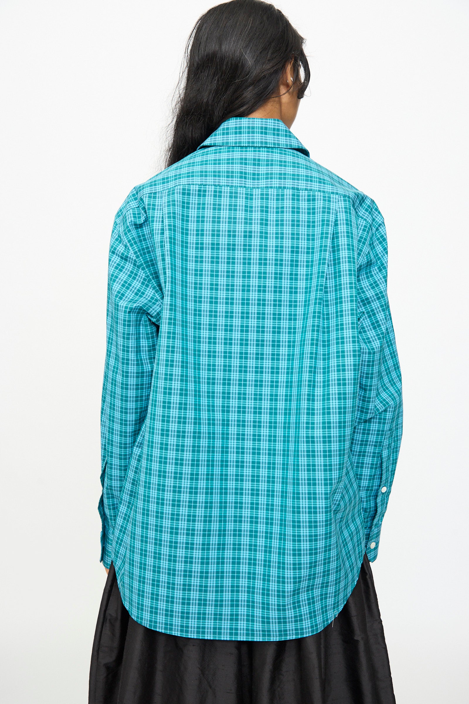 A person with long dark hair faces away, wearing the Olivia Cashmore Rufus Shirt in Blue Green Check, made in New Zealand, paired with a black skirt against a plain white background.