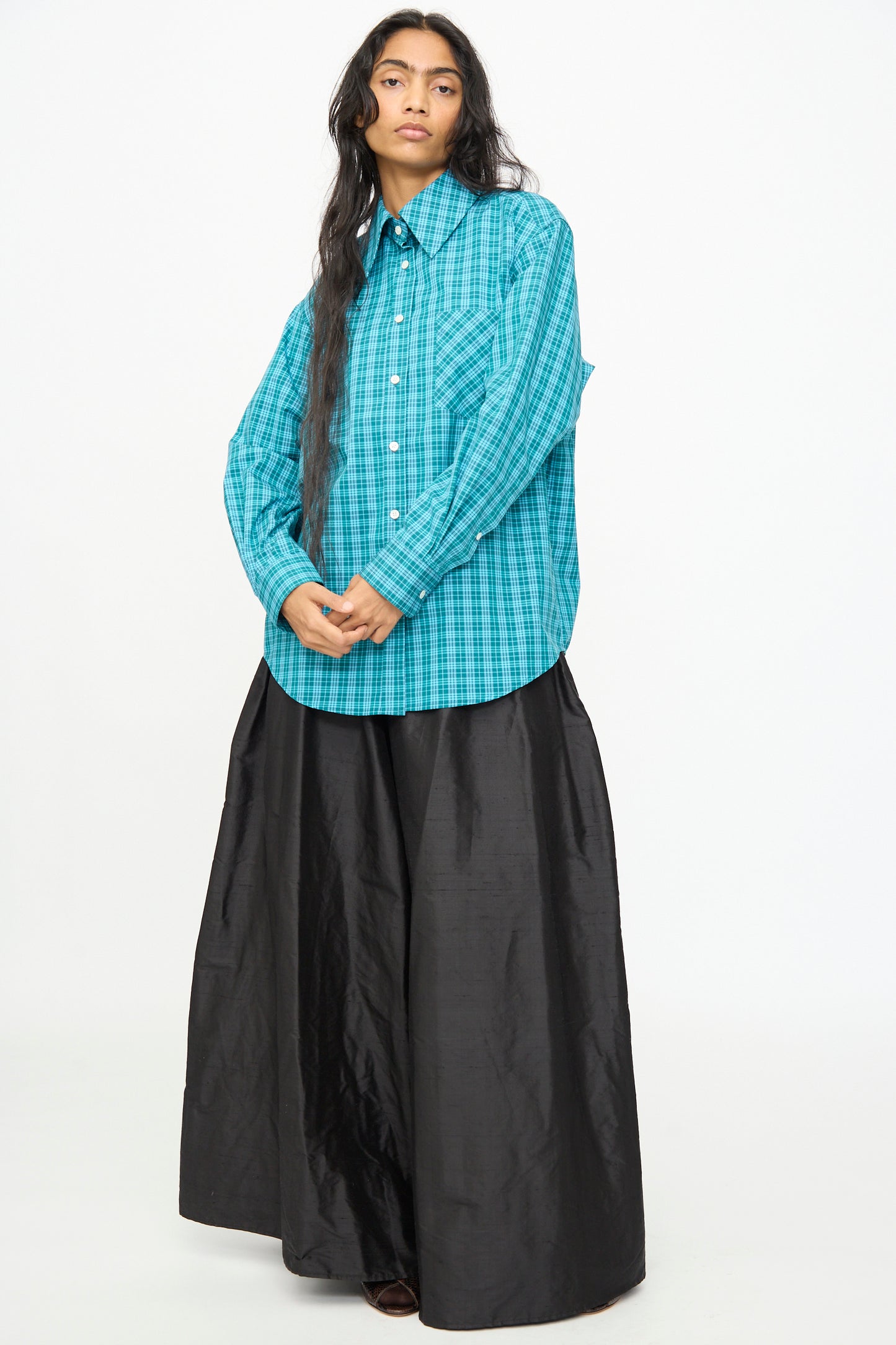 A person wears the Olivia Cashmore Rufus Shirt in Blue Green Check—crafted from cotton in New Zealand—with a long, wide black skirt, standing against a plain white background.