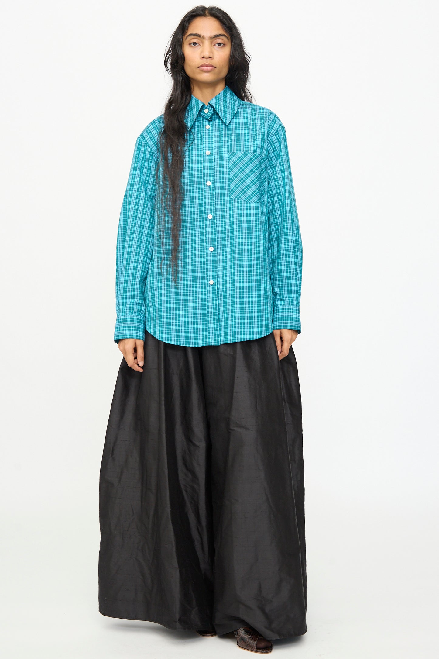 A person stands against a plain background wearing the Olivia Cashmore Rufus Shirt in Blue Green Check and a long, black, wide skirt.