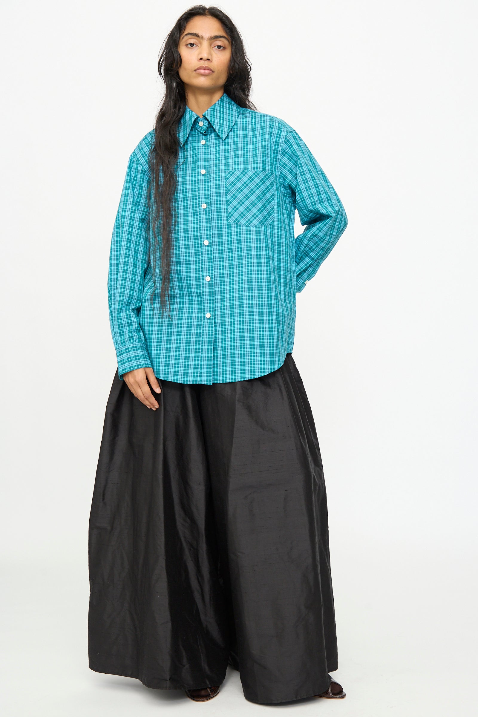 Wearing the Olivia Cashmore Rufus Shirt in Blue Green Check, crafted from cotton with long sleeves and made in New Zealand, the person pairs it with a long, wide black skirt while standing against a plain white background.