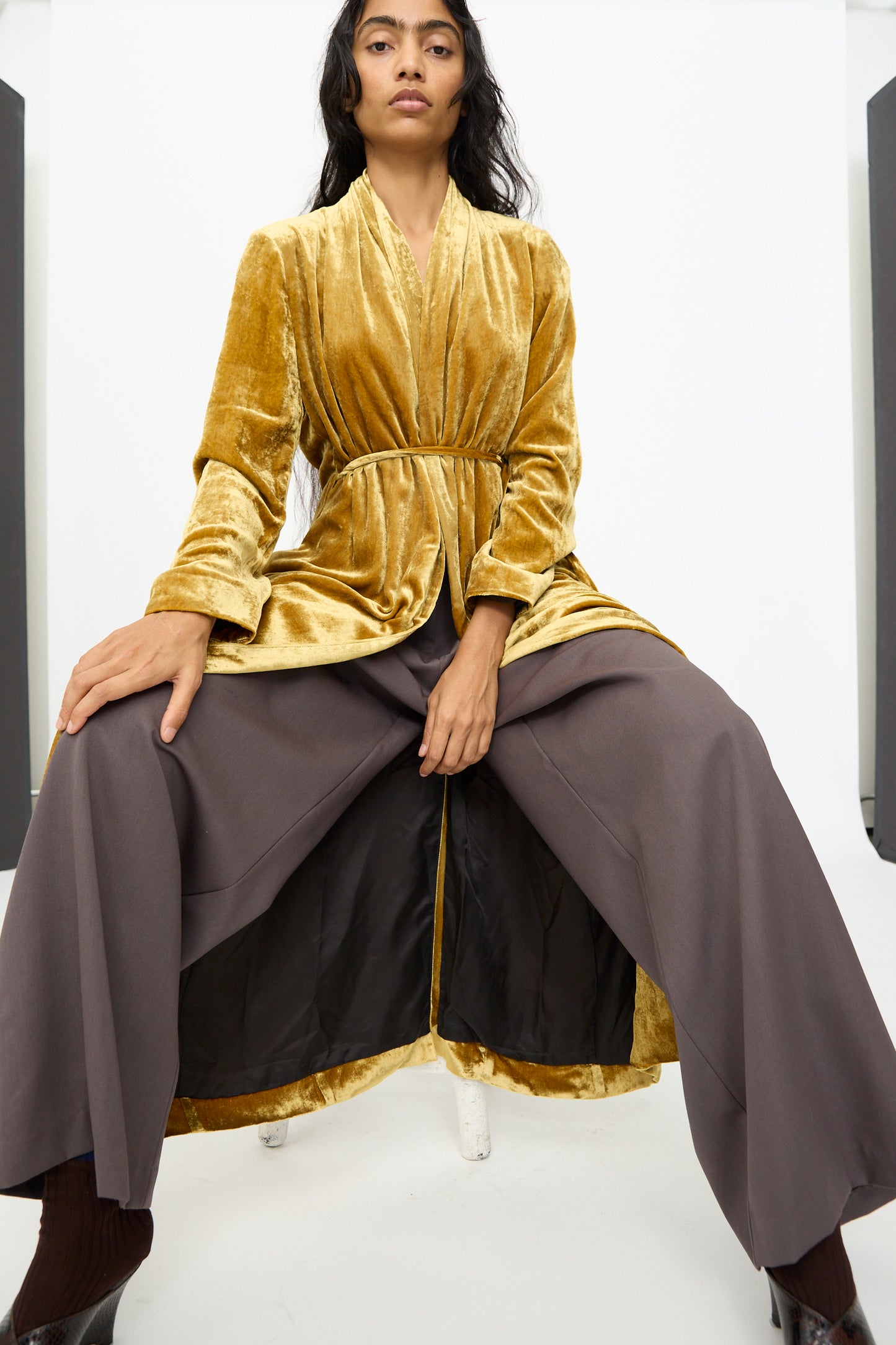 A woman wearing the Olivia Cashmore Theresa Coat in Amber, belted at the waist over wide-legged gray pants, sits on a white chair against a plain background.