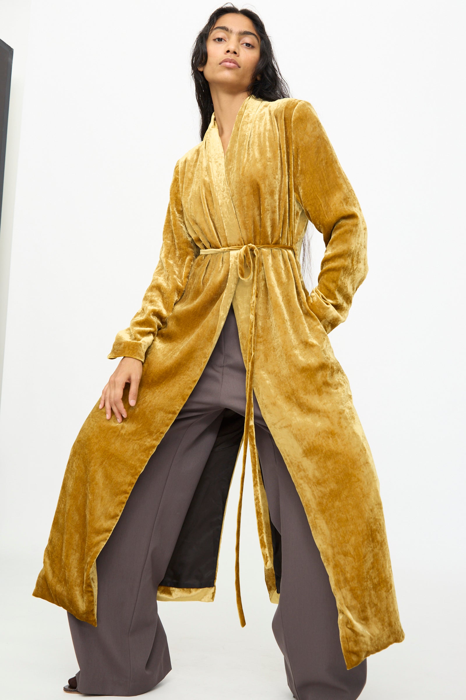 Wearing the Olivia Cashmore Theresa Coat in Amber—a long, gold velvet wrap coat with a tie belt—paired with wide-legged brown pants, standing against a plain white background.
