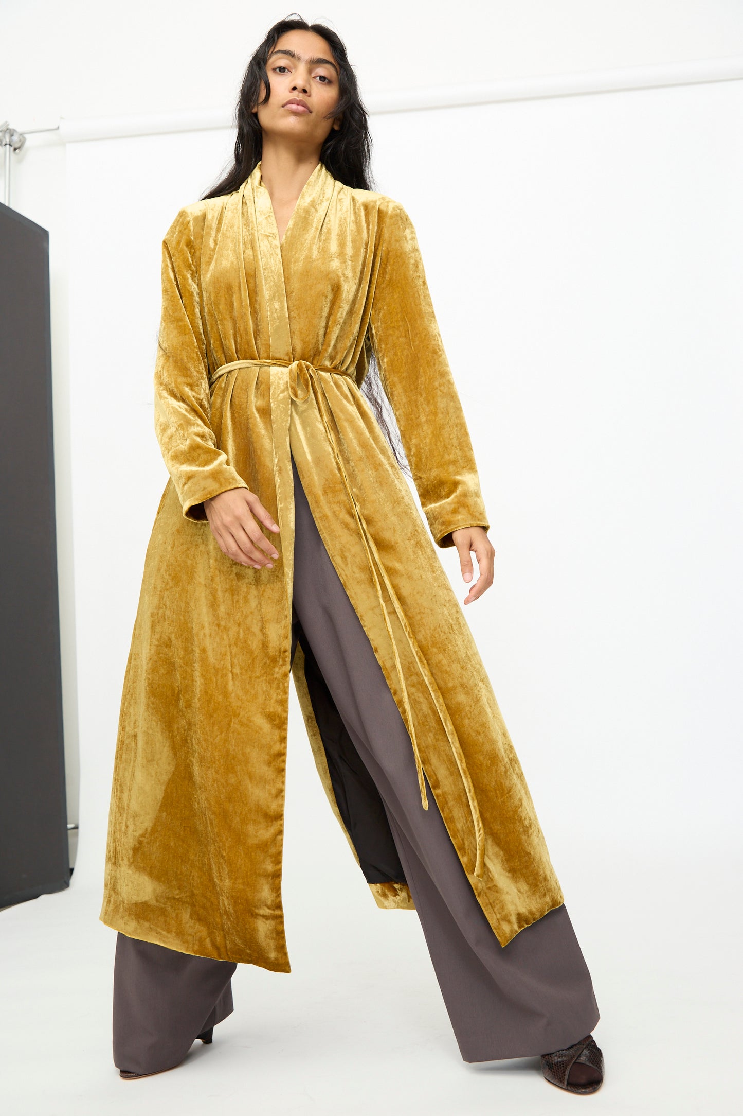 Wearing the Olivia Cashmore Theresa Coat in Amber—a luxurious, long viscose silk design cinched at the waist—over wide-leg brown pants, a person stands against a plain studio backdrop.