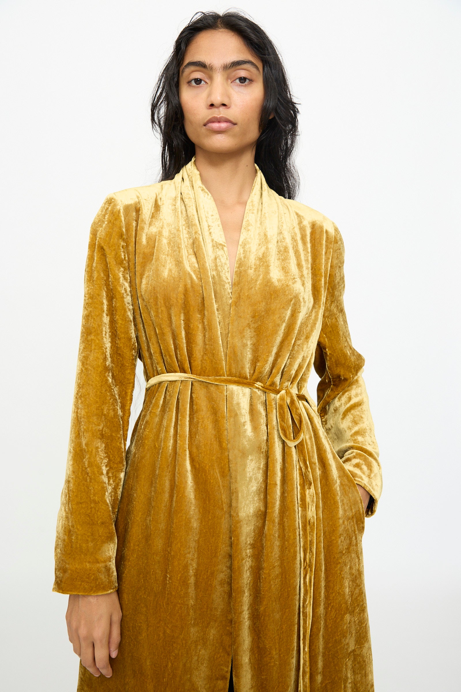 A person wearing the Theresa Coat in Amber by Olivia Cashmore, a gold velvet belted robe-style wraparound coat, stands against a plain white background.