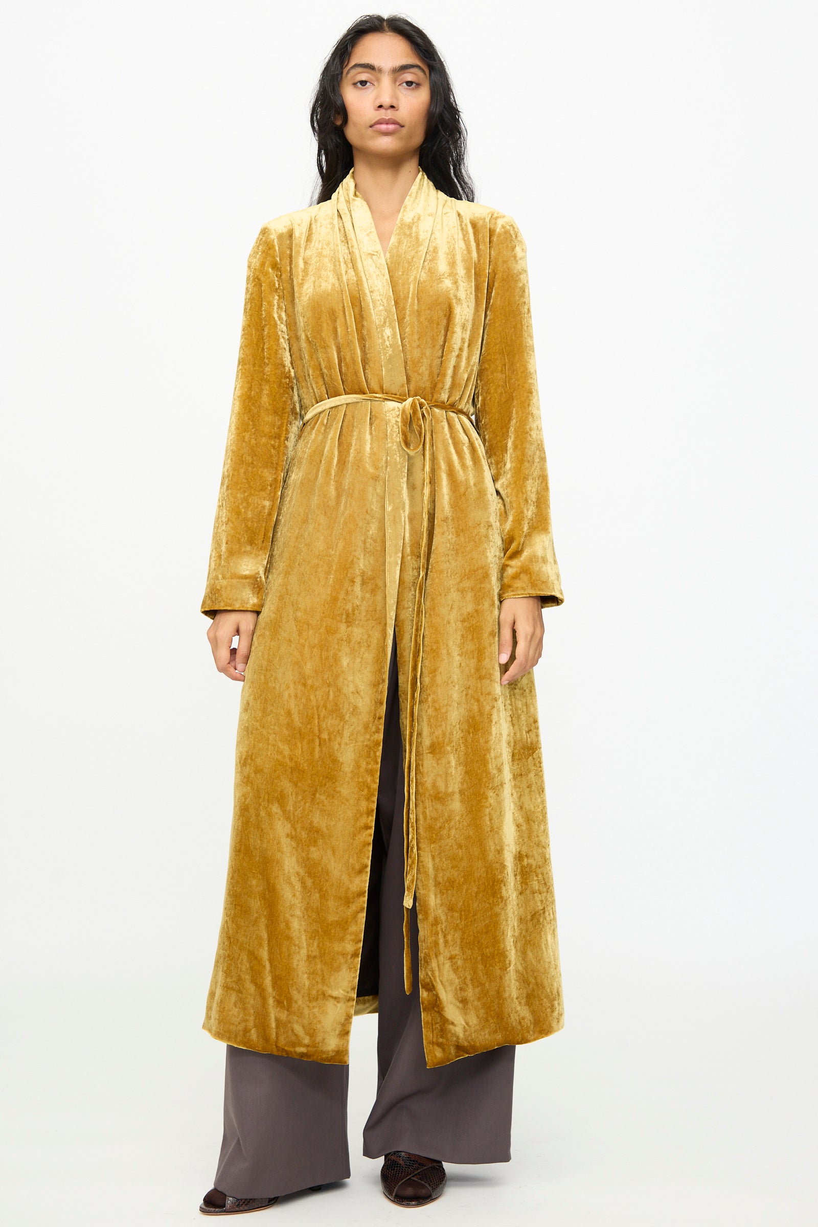 A model stands facing forward against a plain white background, wearing the Olivia Cashmore Theresa Coat in Amber, styled as a belted robe over wide-legged brown pants.