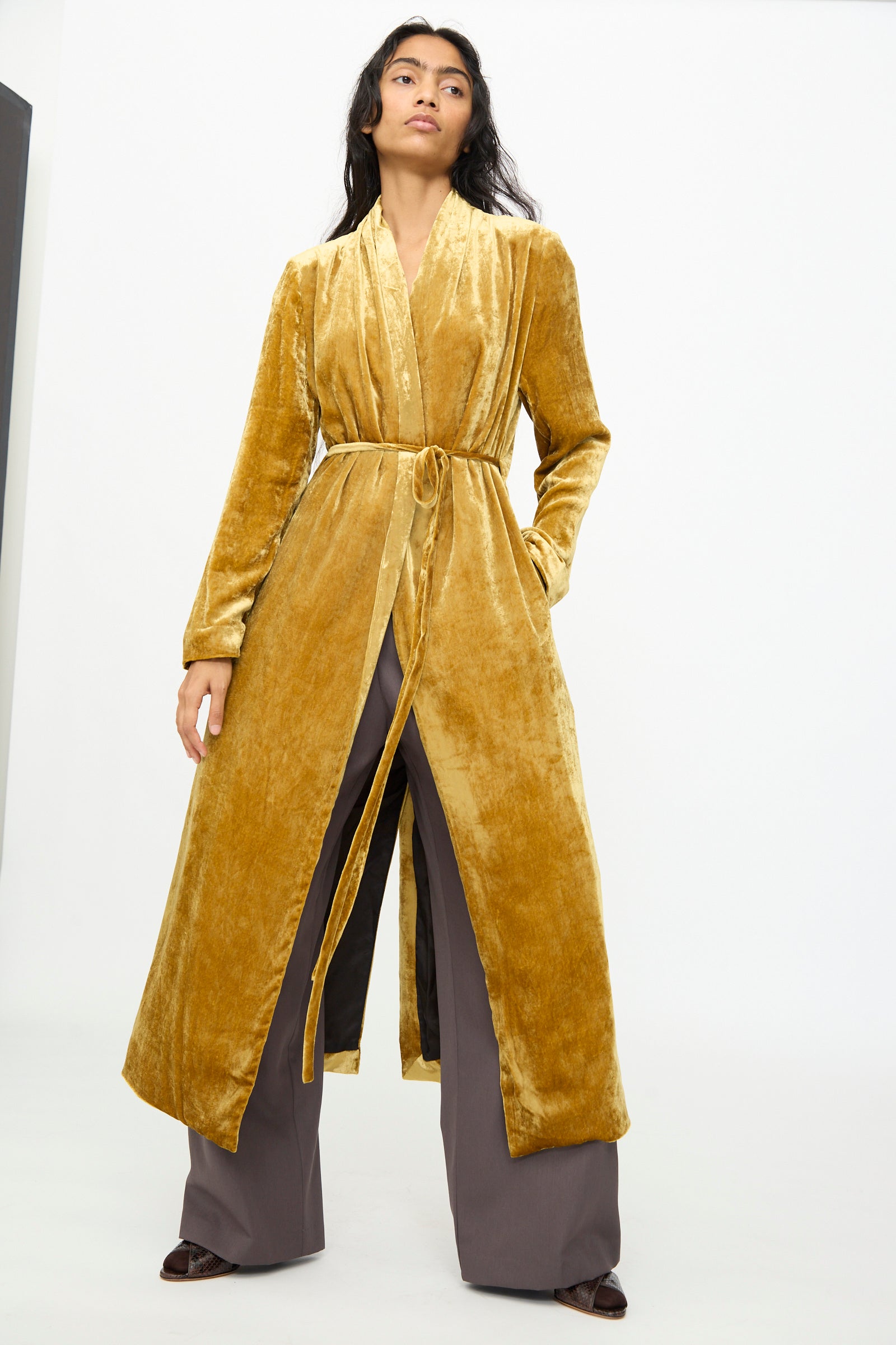 A woman stands against a plain white background wearing the Olivia Cashmore Theresa Coat in Amber, styled over wide-leg gray pants and brown shoes.