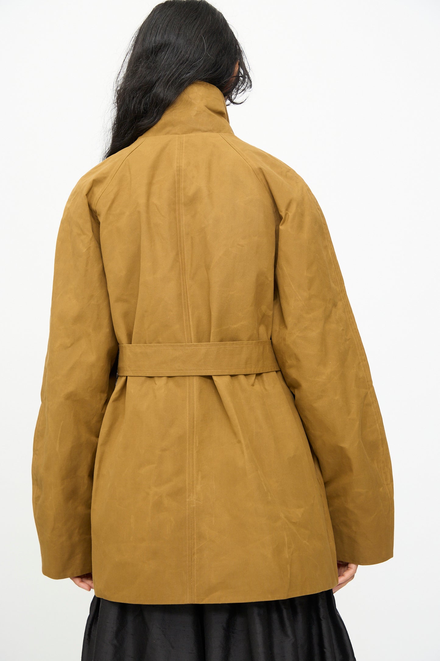 A person with long dark hair is seen from the back, wearing the Olivia Cashmore Waxed Cotton Morison Parka in Ochre, crafted from waxed cotton and made in New Zealand, layered over a black skirt against a plain background.
