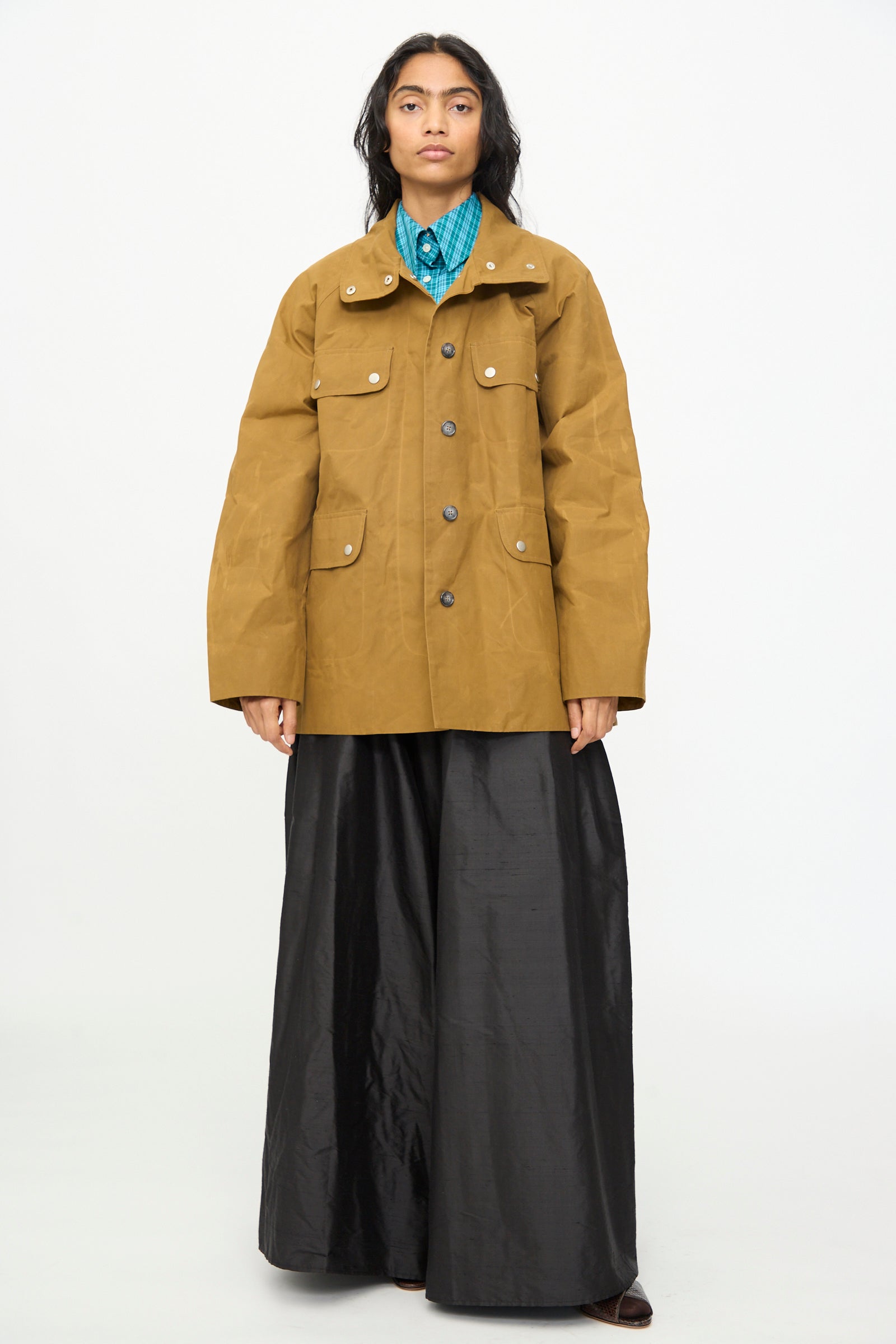 A person stands against a plain background, wearing an Olivia Cashmore Waxed Cotton Morison Parka in Ochre, made in New Zealand, over a turquoise shirt and wide black satin pants.